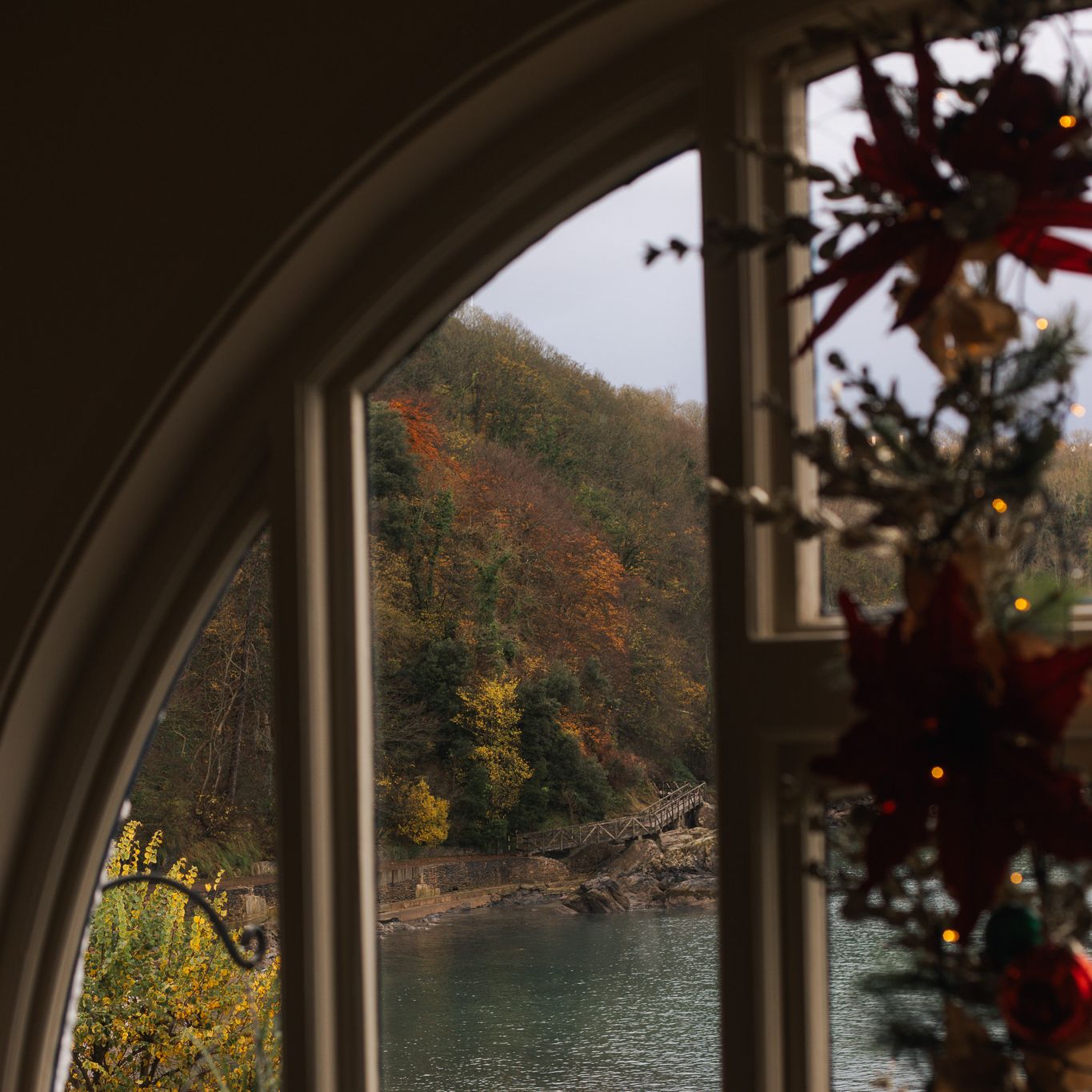 View through an arched window showing a scenic riverside with autumn foliage and a decorated Christmas tree inside.