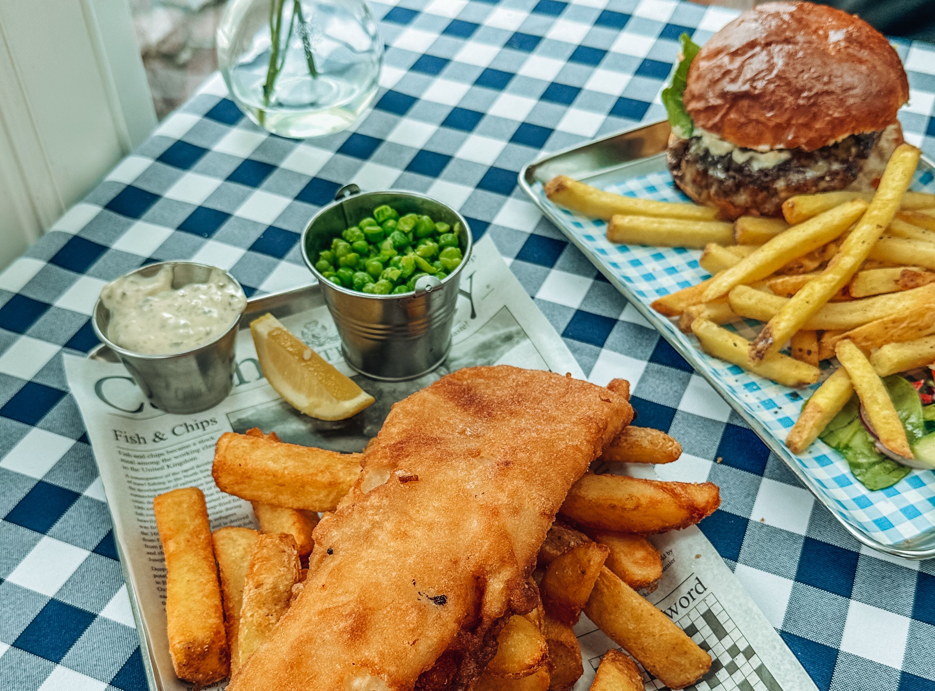 Fish and chips served with tartar sauce, peas, and lemon wedge on a checkered tablecloth, with a burger and fries in the background.