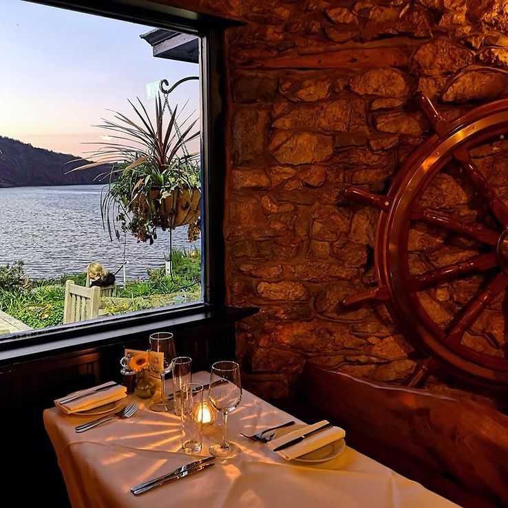 Cozy stone-walled restaurant with nautical decor, featuring a wooden ship's wheel and a set dinner table by a window overlooking a scenic waterfront at sunset.