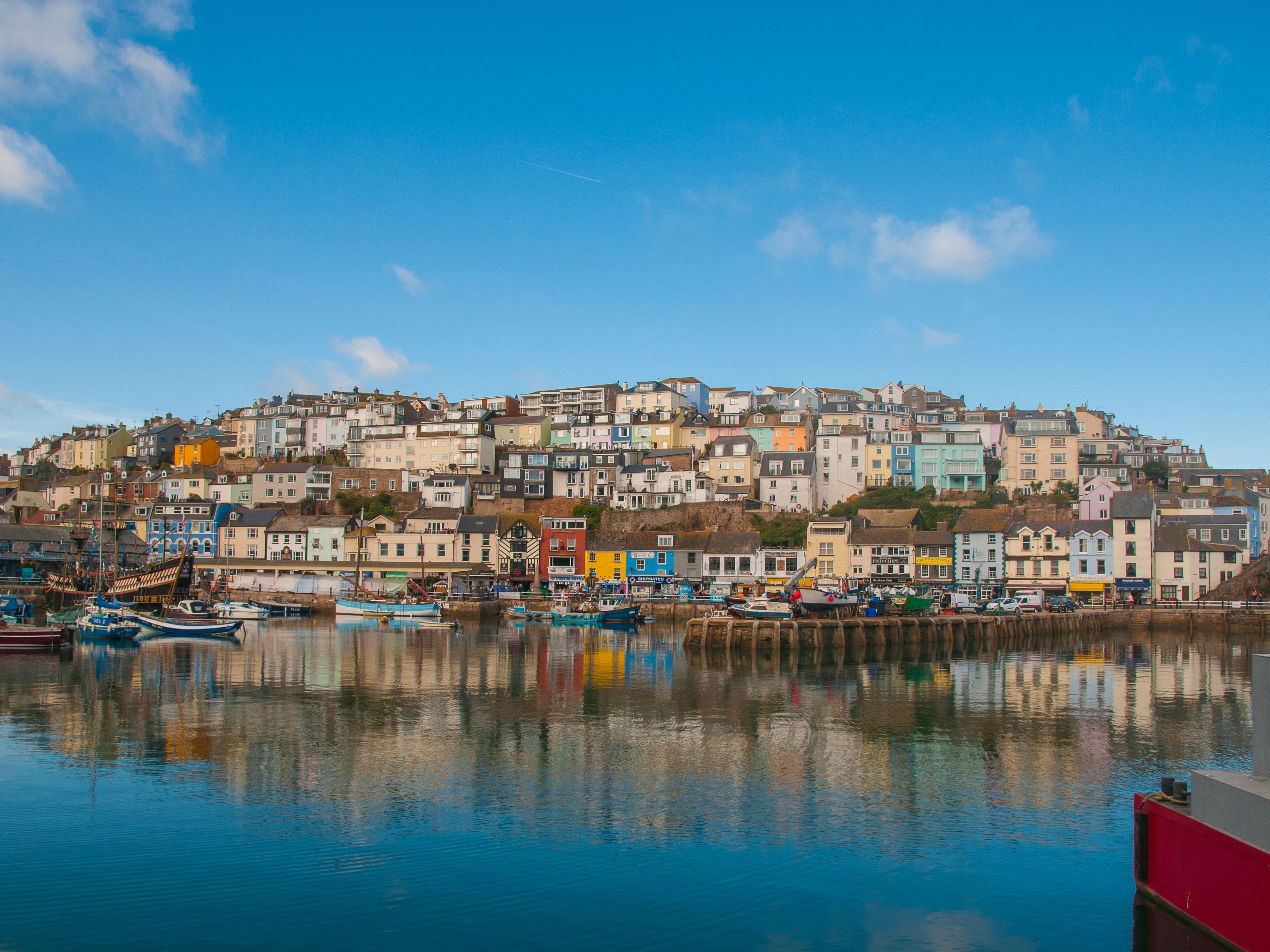 Colorful houses on a hillside overlooking a harbor with boats, reflected in the water under a blue sky.