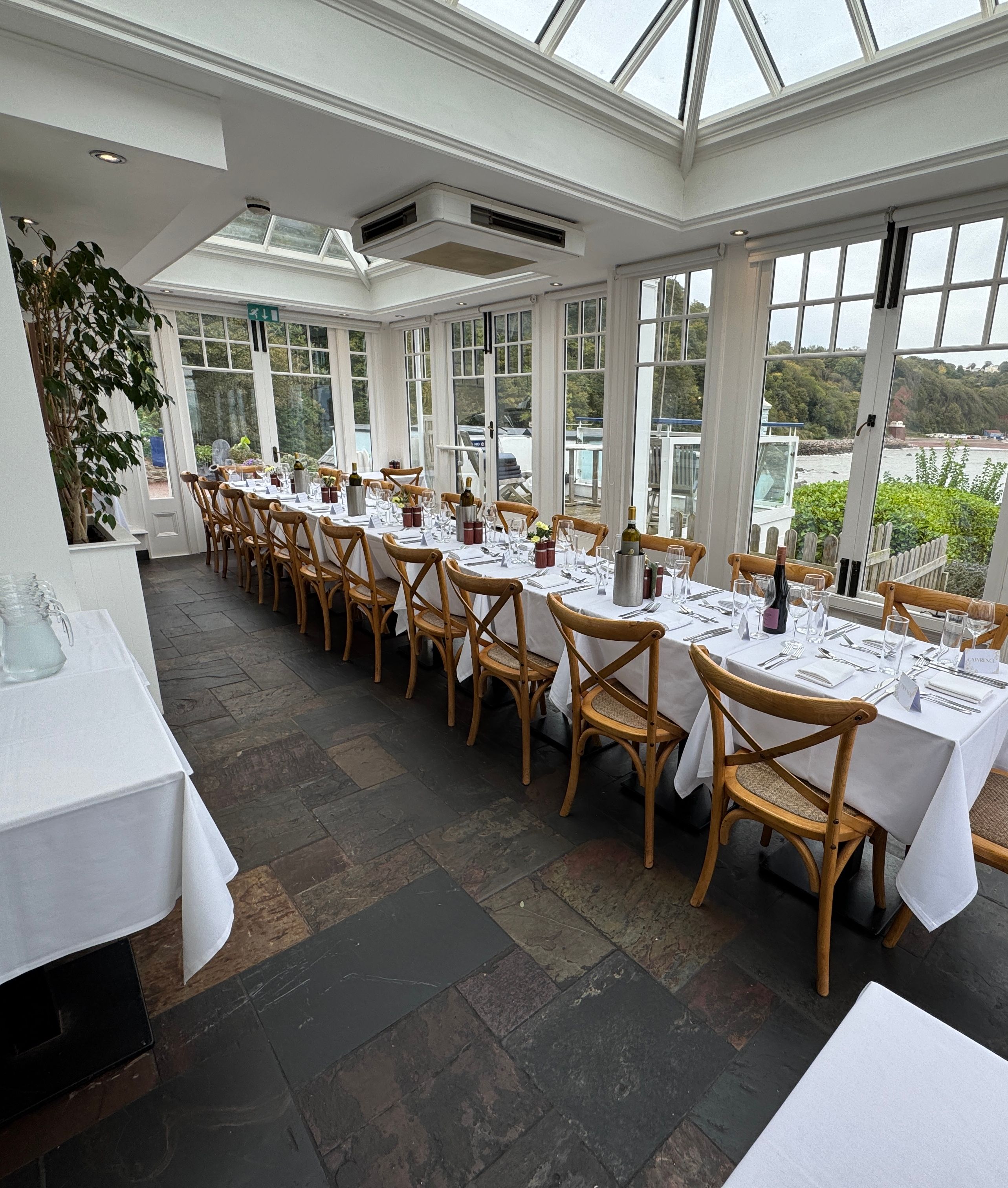 Elegant dining area set for a meal in a bright conservatory with large windows overlooking a river and greenery.