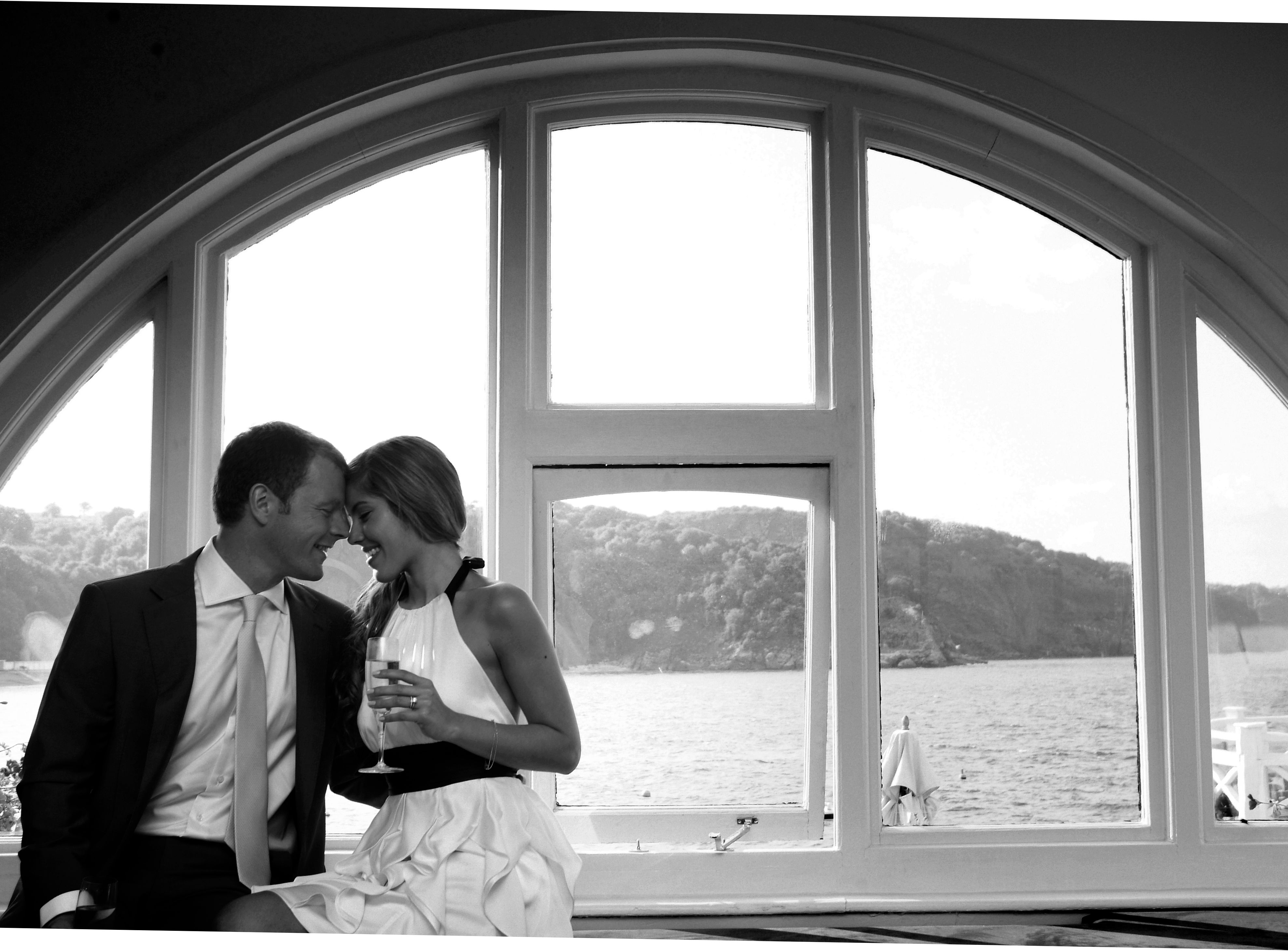 A couple sitting closely together by a large arched window, overlooking a scenic waterfront view.