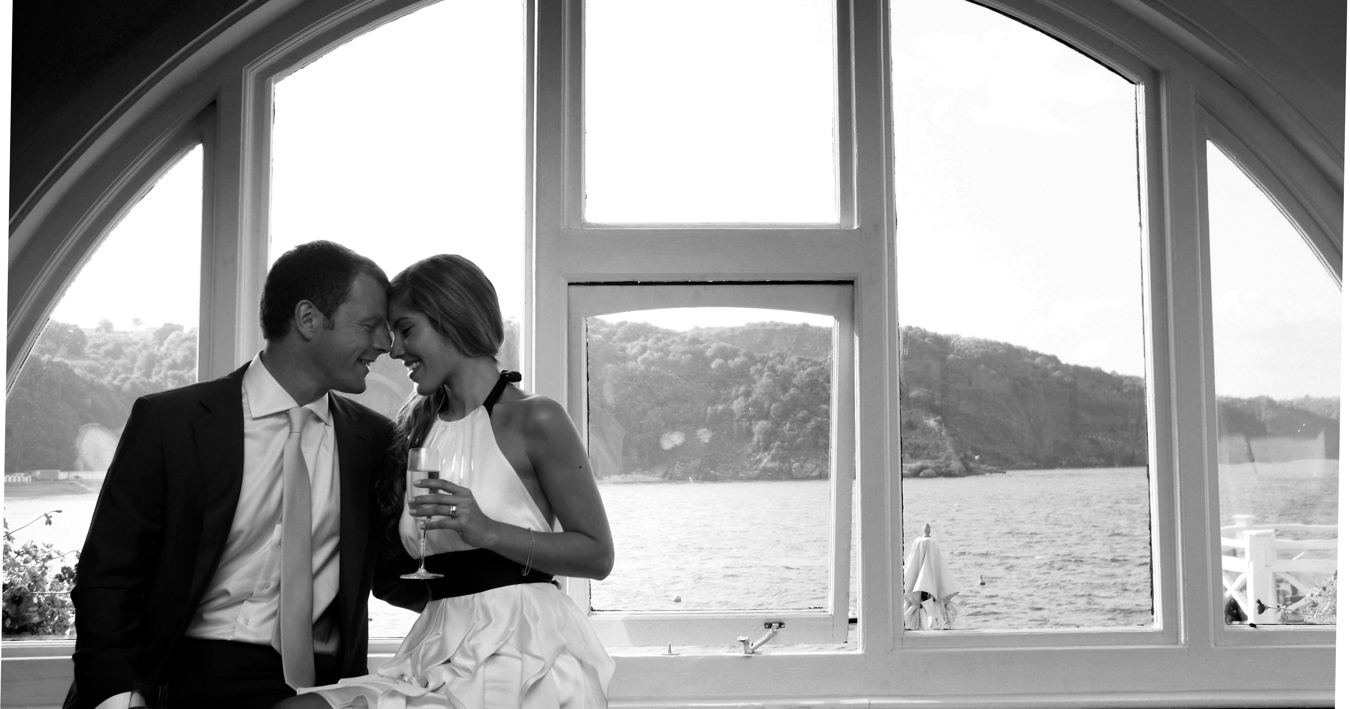 A couple sitting closely together by a large arched window, overlooking a scenic waterfront view.