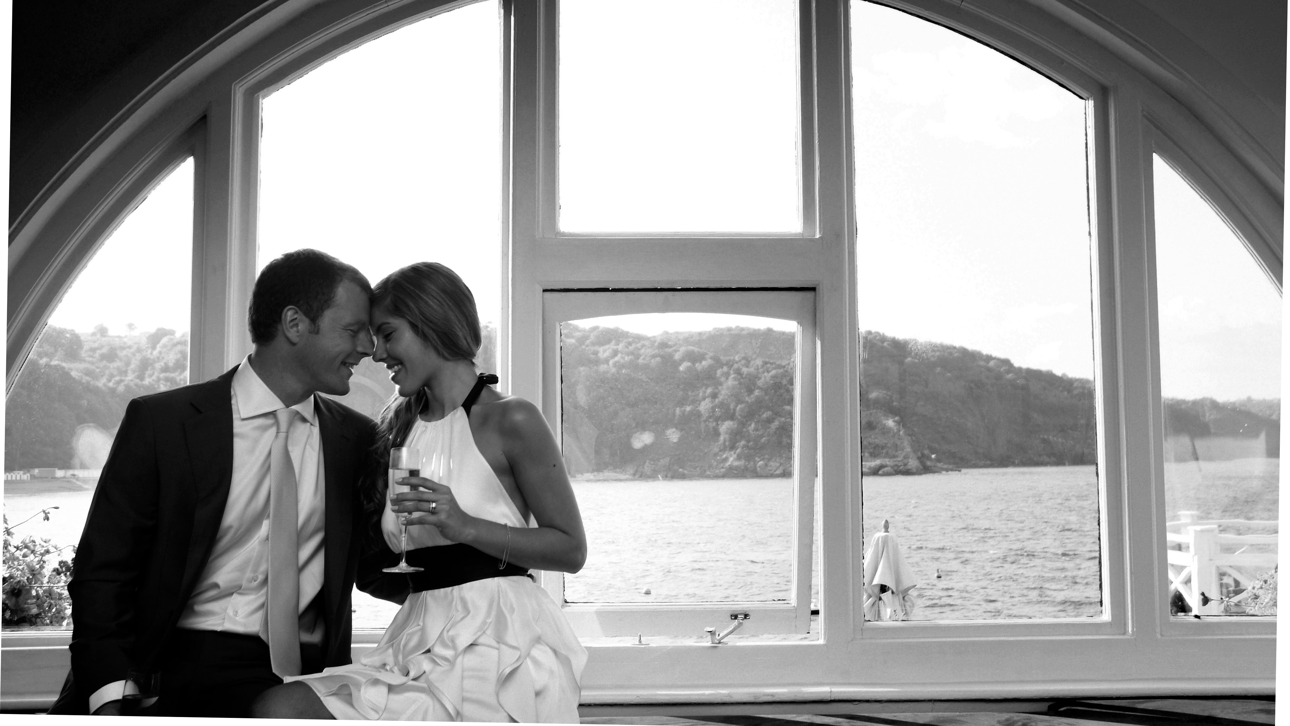 A couple sitting closely together by a large arched window, overlooking a scenic waterfront view.