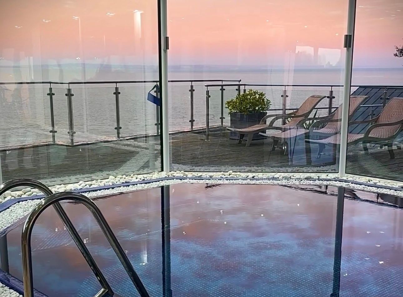 Indoor swimming pool with glass walls overlooking a balcony with lounge chairs and a scenic ocean view at sunset.