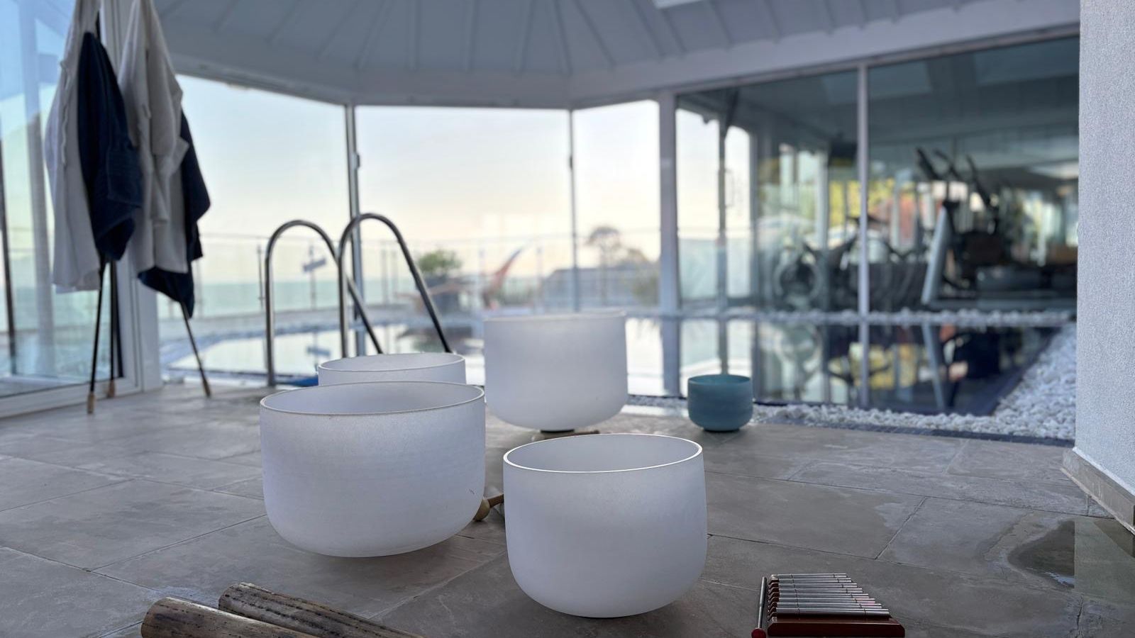 Crystal singing bowls and mallets set up near an indoor pool with a view of a gym through glass walls.