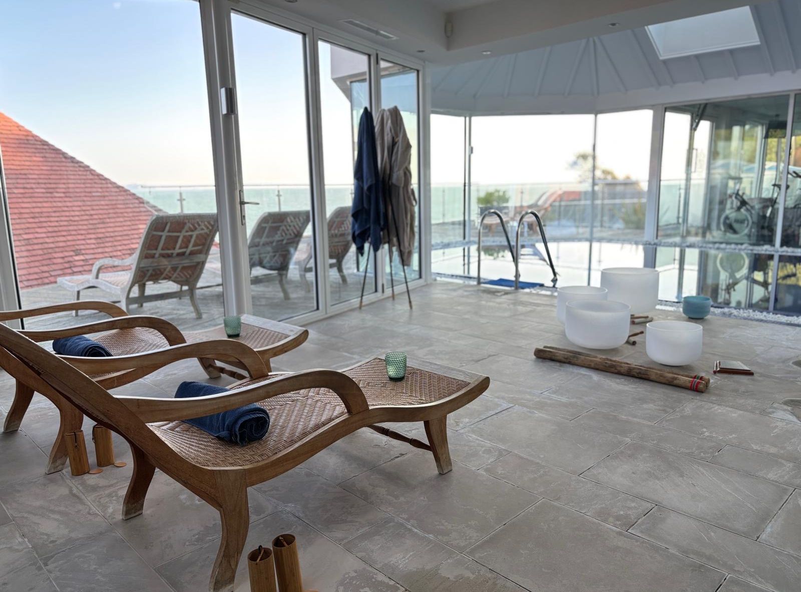 Indoor spa area with lounge chairs, a pool, and glass walls overlooking an outdoor terrace and ocean view.