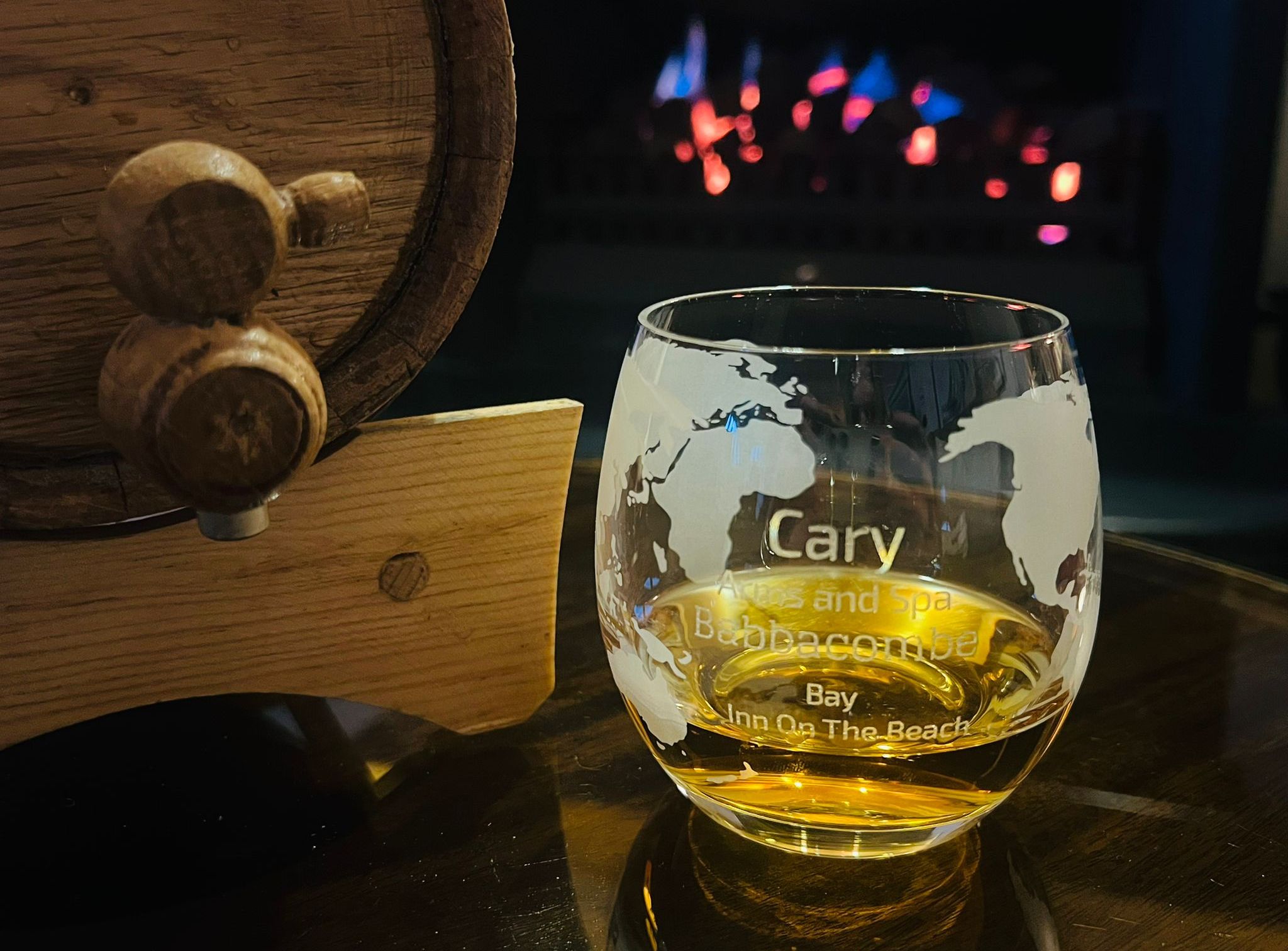 A glass of whiskey with engraved text next to a small wooden barrel, with a fireplace in the background.