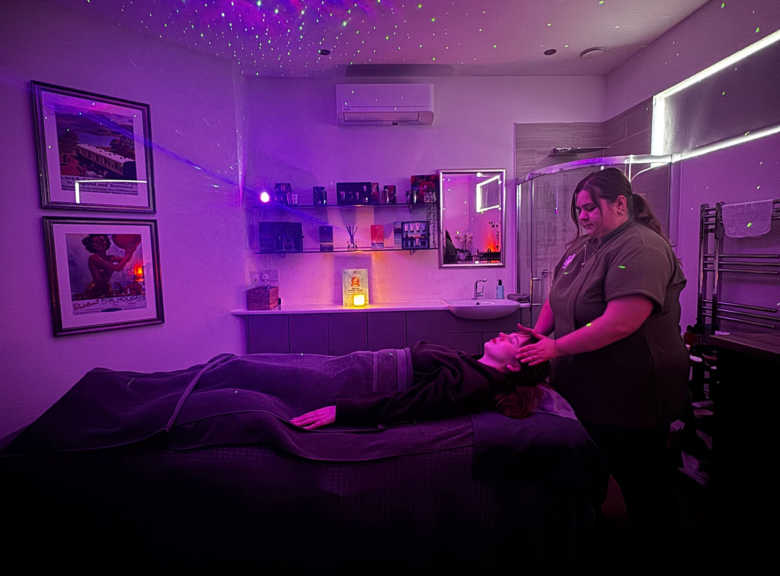 Therapist giving a head massage to a client on a treatment bed in a spa room lit with purple galaxy-style lights.
