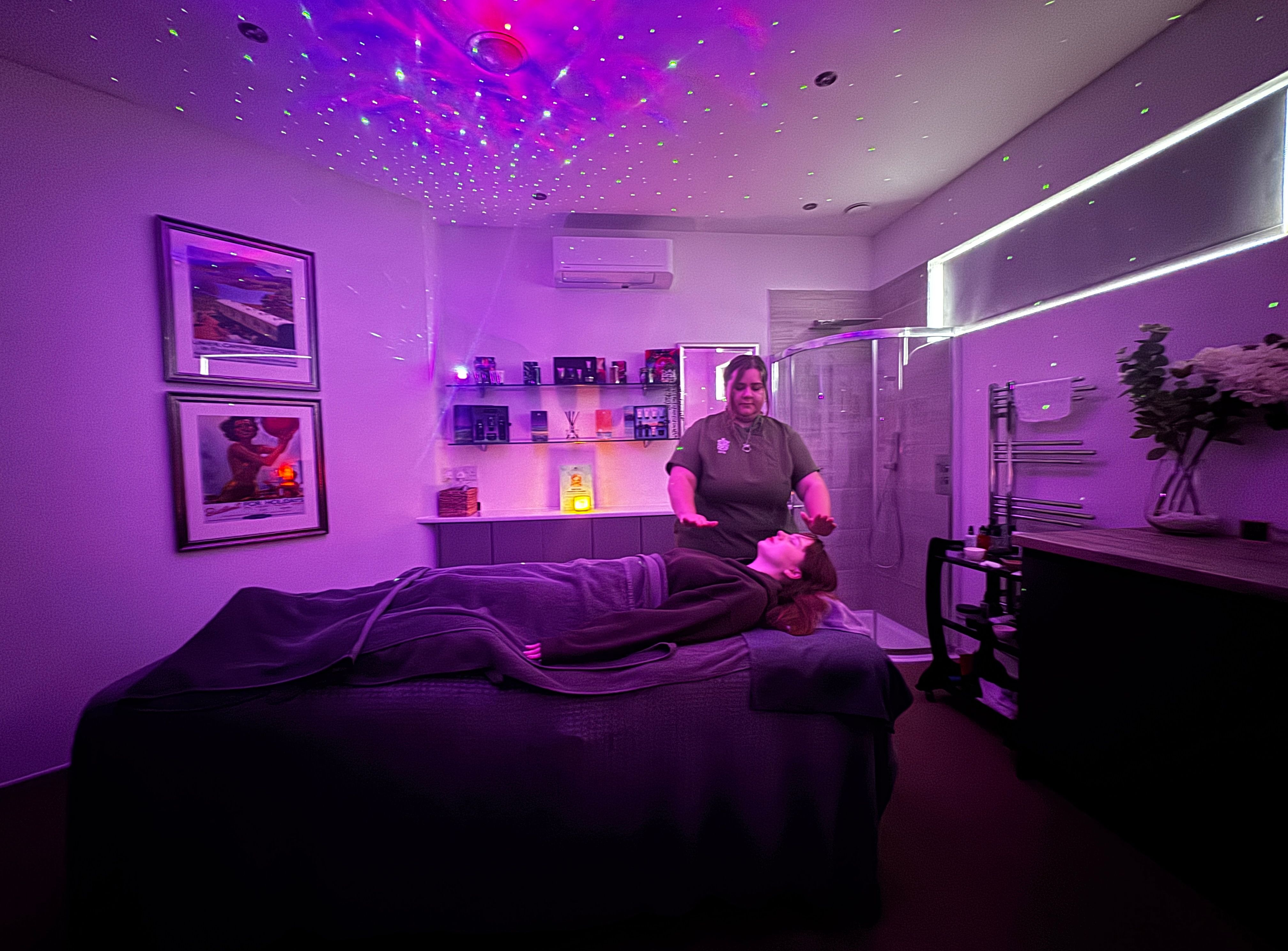 Therapist performing a reiki-style energy healing treatment on a client lying on a massage table under purple starry projector lights in a spa room.
