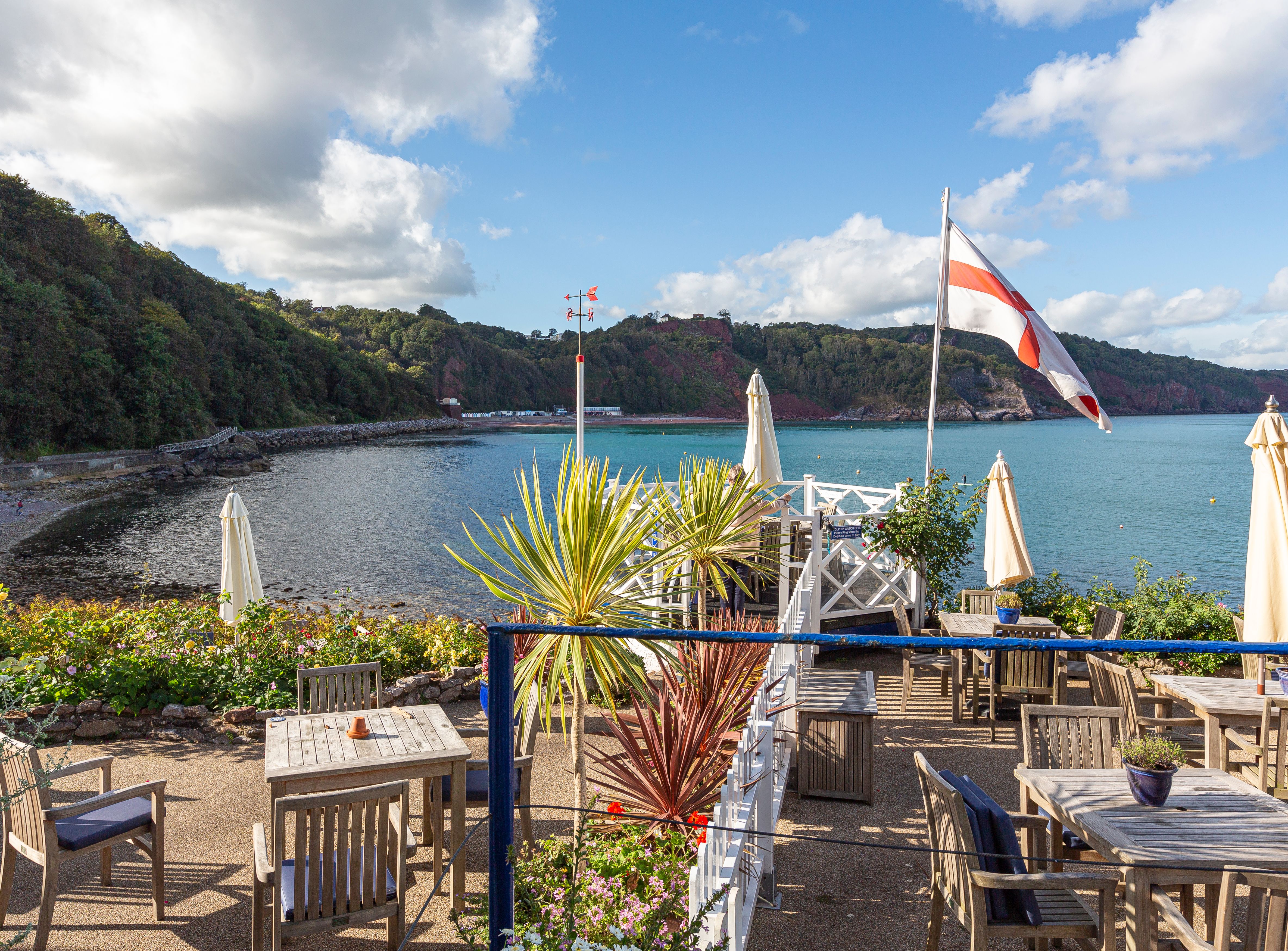 Outdoor terrace with wooden tables, parasols and an England flag overlooking a calm coastal bay