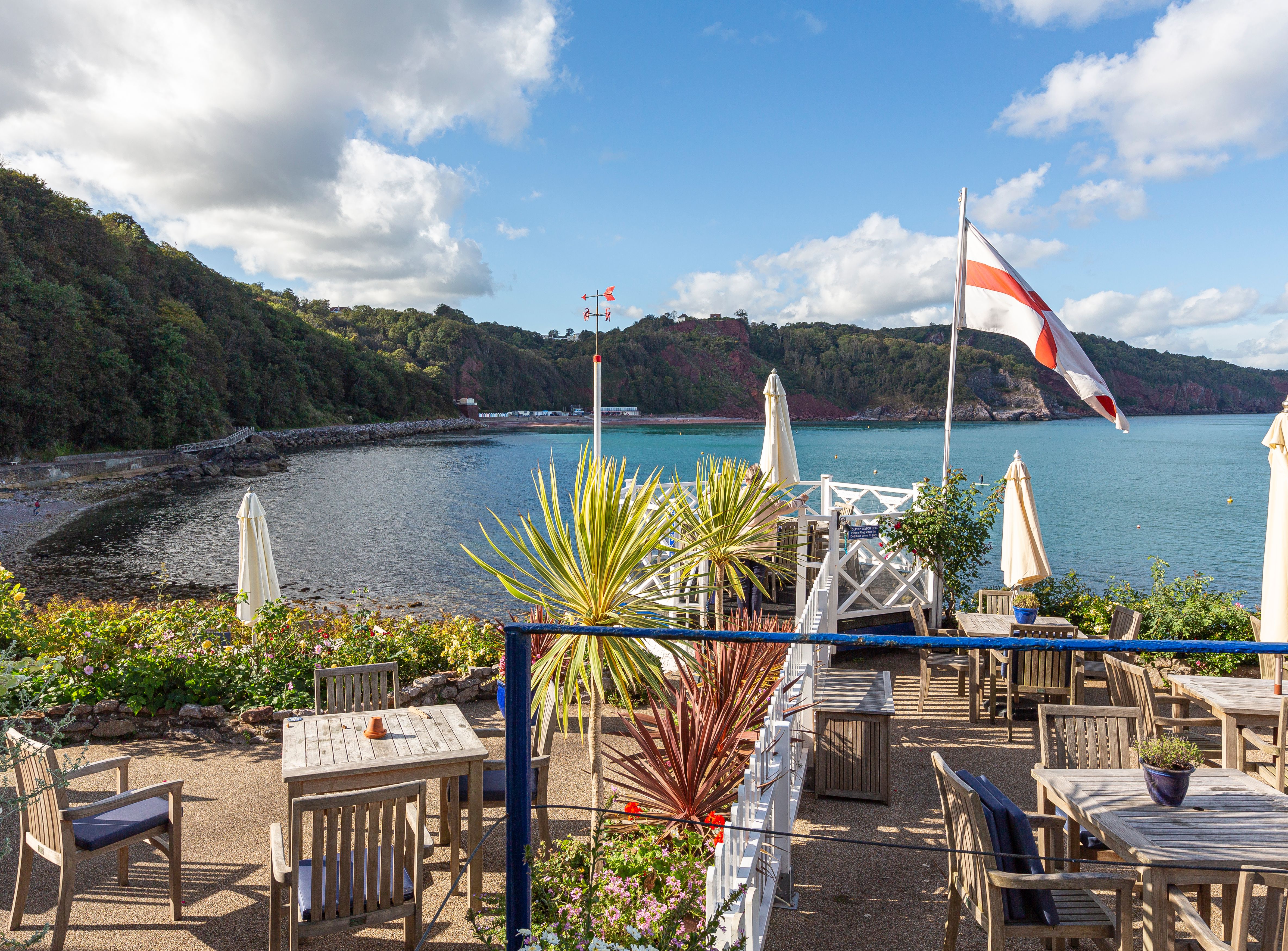 Outdoor terrace with wooden tables, parasols and an England flag overlooking a calm coastal bay