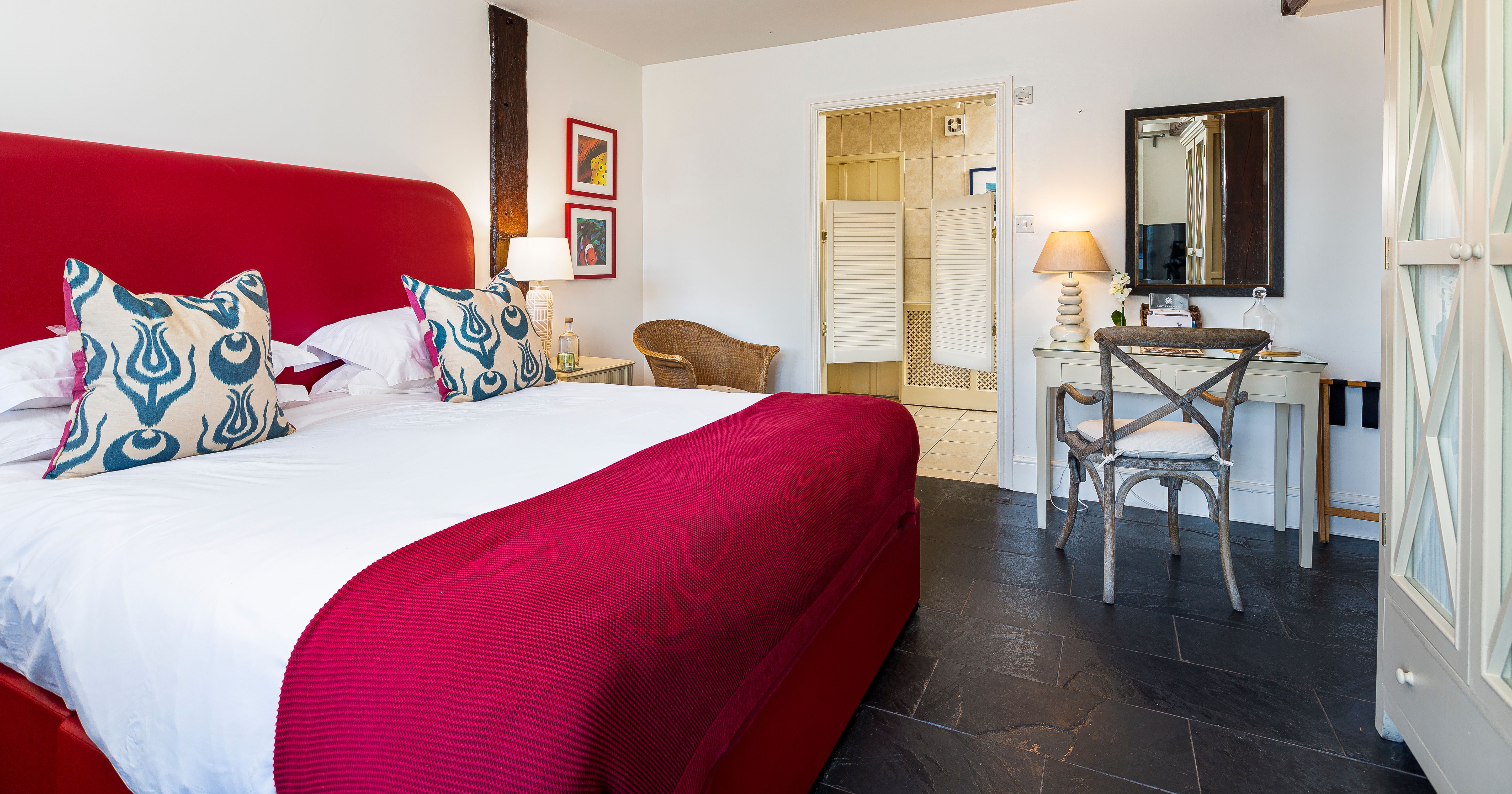 Bright bedroom with red upholstered bed, patterned cushions, writing desk and open doorway to en-suite