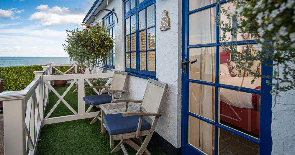 Seaside cottage terrace with blue-framed windows, wooden chairs and ocean view