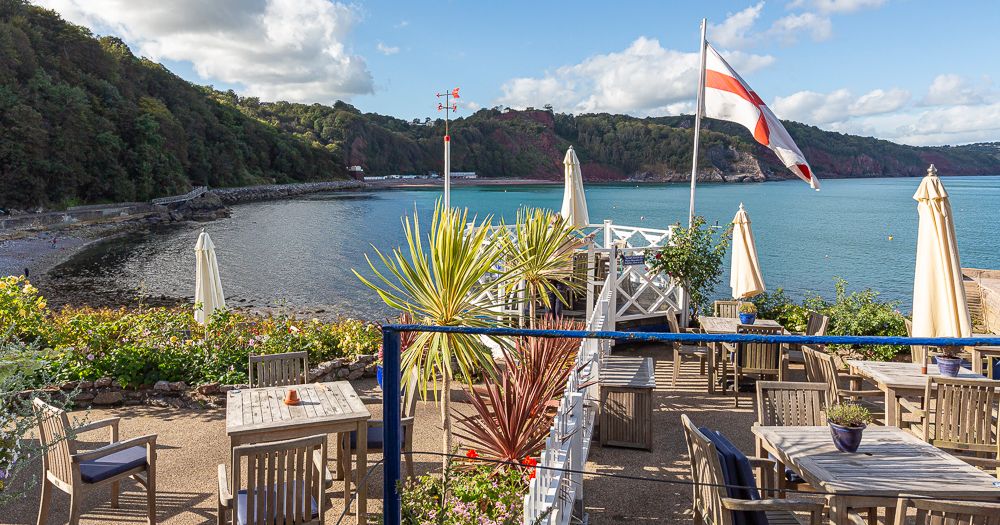 Seaside terrace with wooden tables and umbrellas overlooking a calm bay, with a red and white flag flying under a blue sky