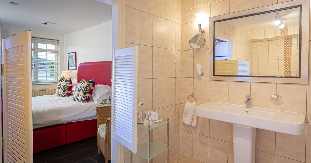 Tiled bathroom with pedestal sink and large mirror, opening to a bedroom with a red headboard
