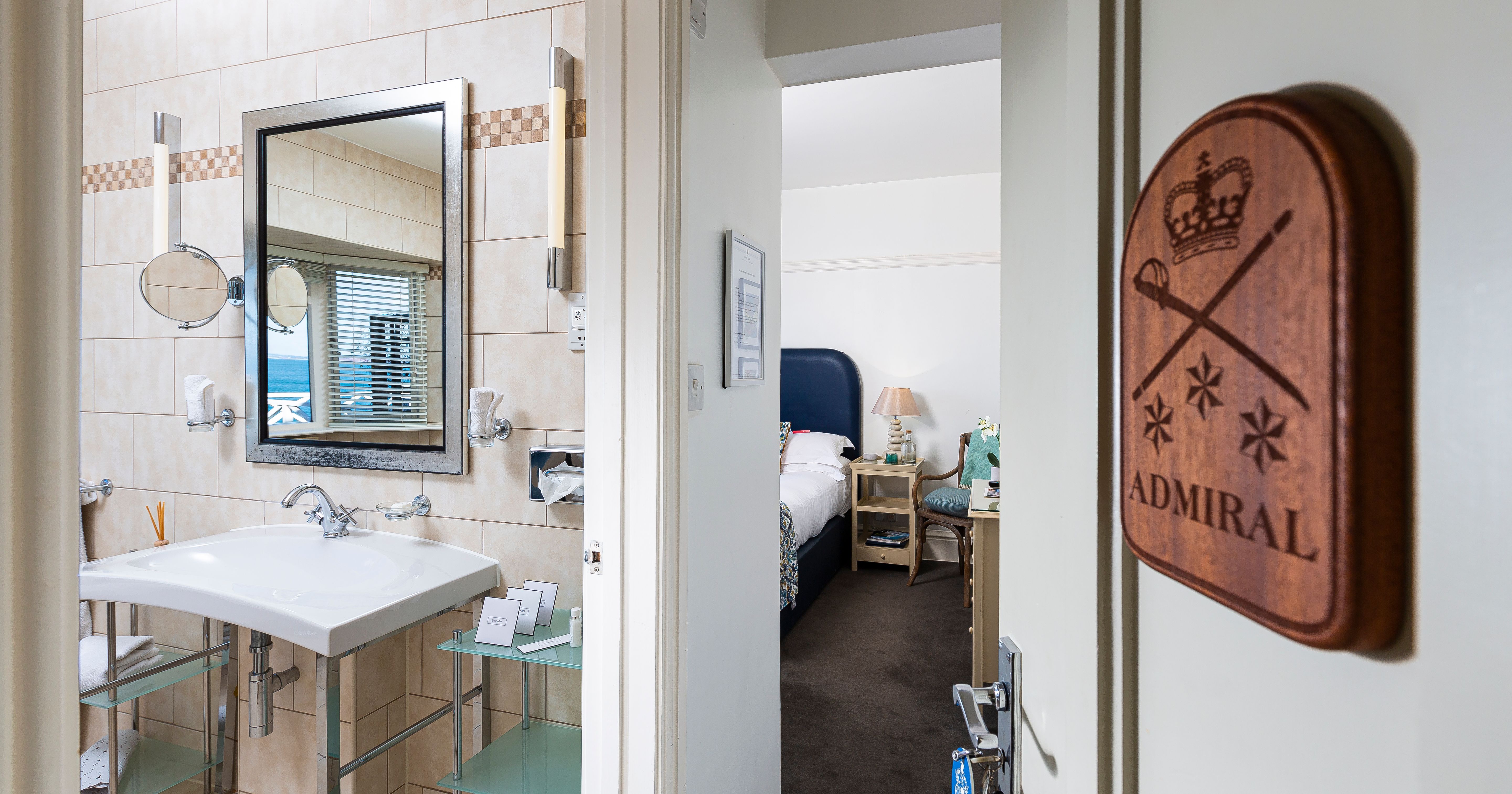View through an open door showing a tiled bathroom with sink and mirror beside a bedroom, with an 
