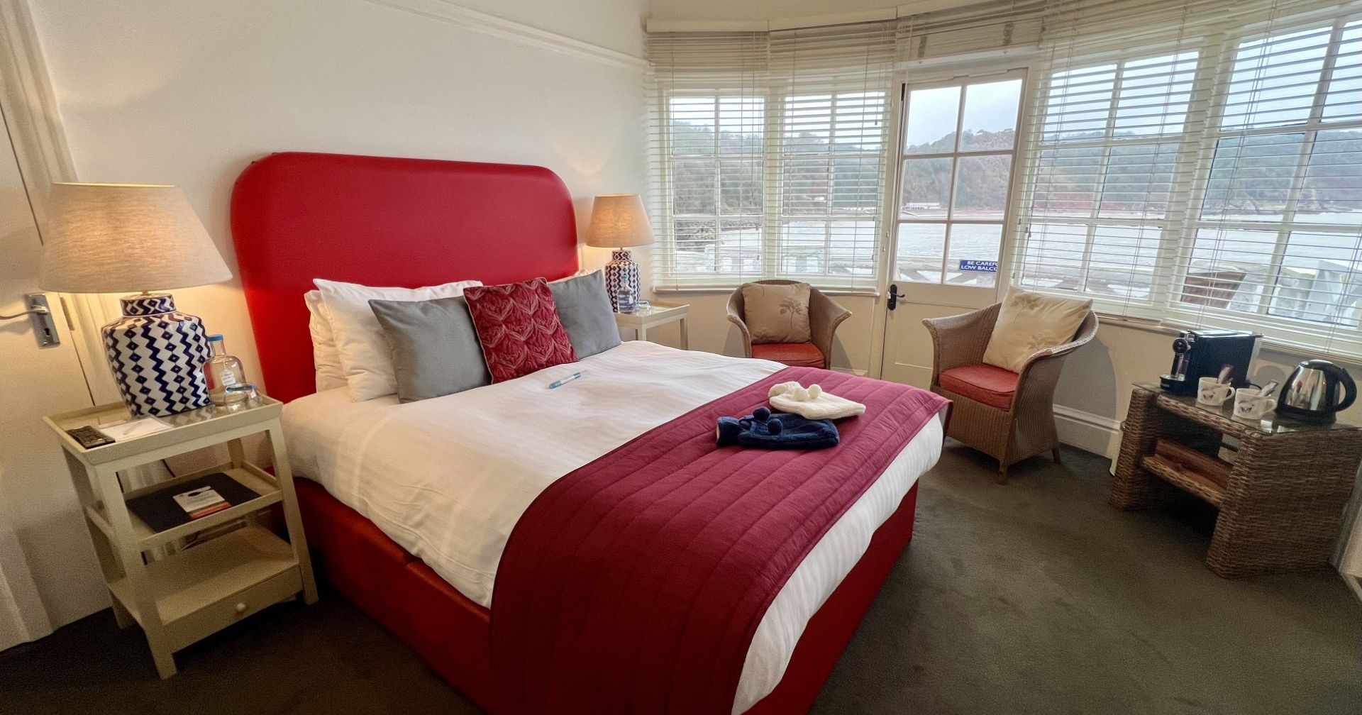 Bright bedroom with red bed frame, white bedding and bay window overlooking water