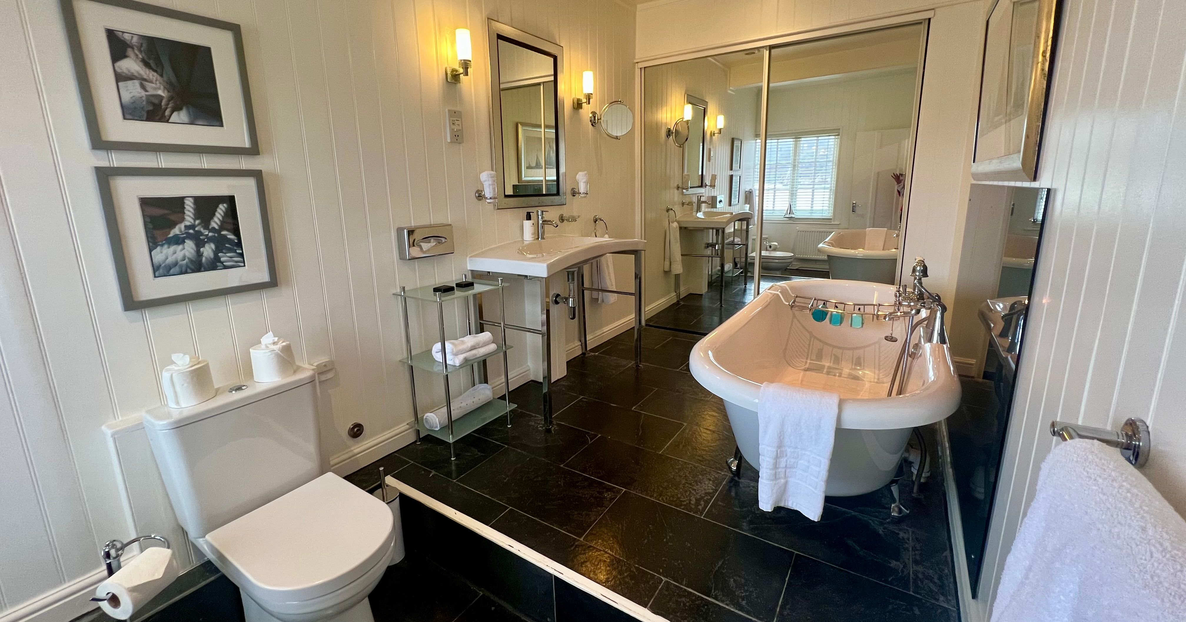 Bright bathroom with freestanding roll-top bath, pedestal sink, toilet and black tiled floor