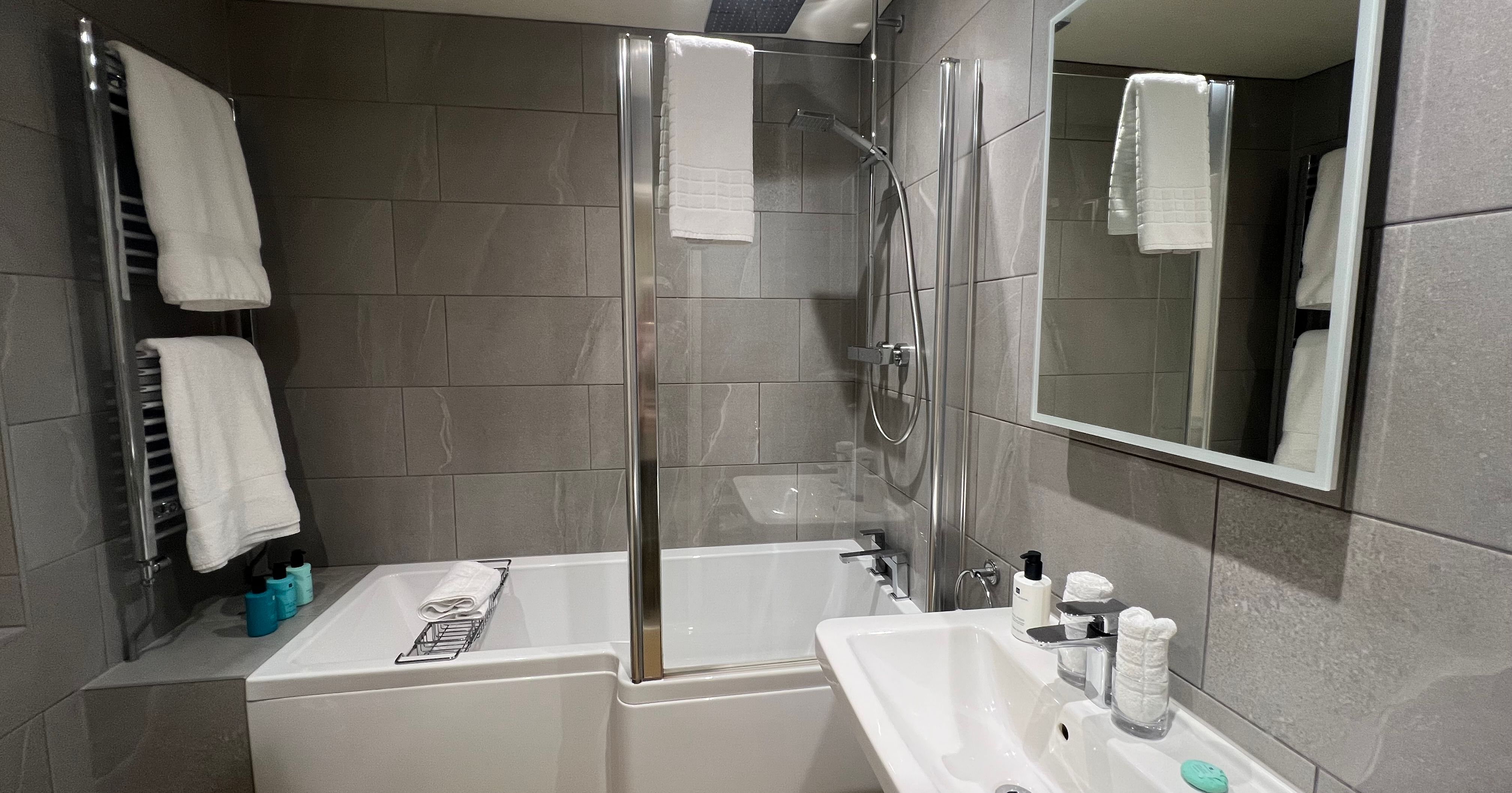 Modern grey-tiled bathroom with bath and shower screen, sink and mirror, and white towels on a heated rail.