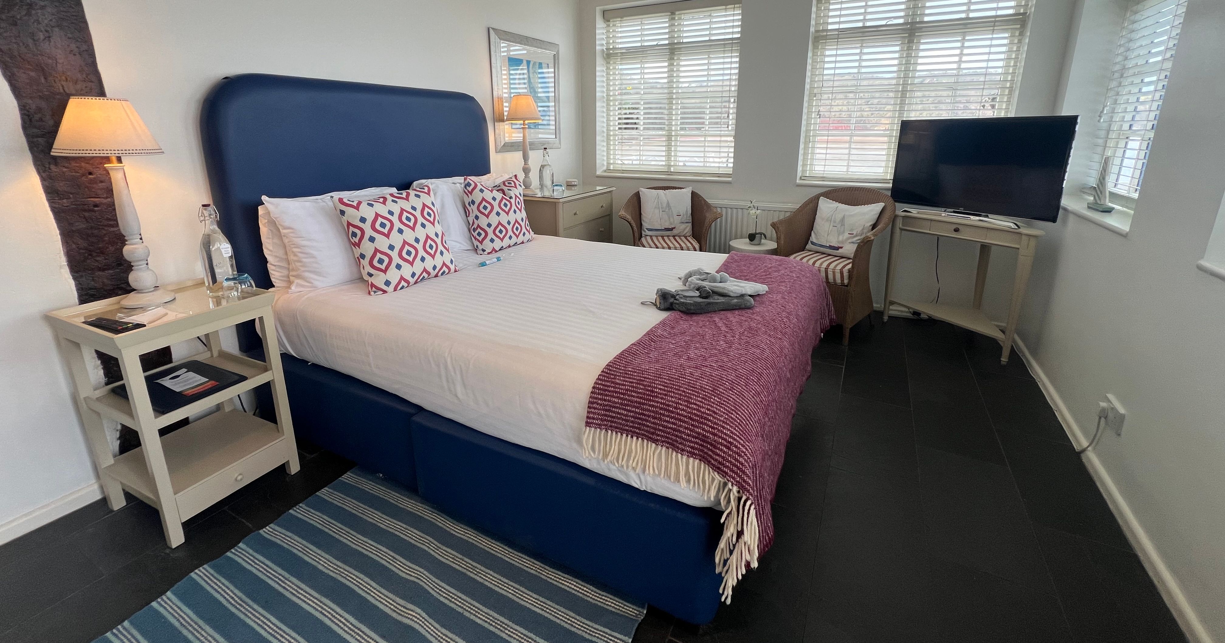 Bright bedroom with blue upholstered bed, patterned cushions, purple throw, bedside lamps, armchairs and a TV by shuttered windows