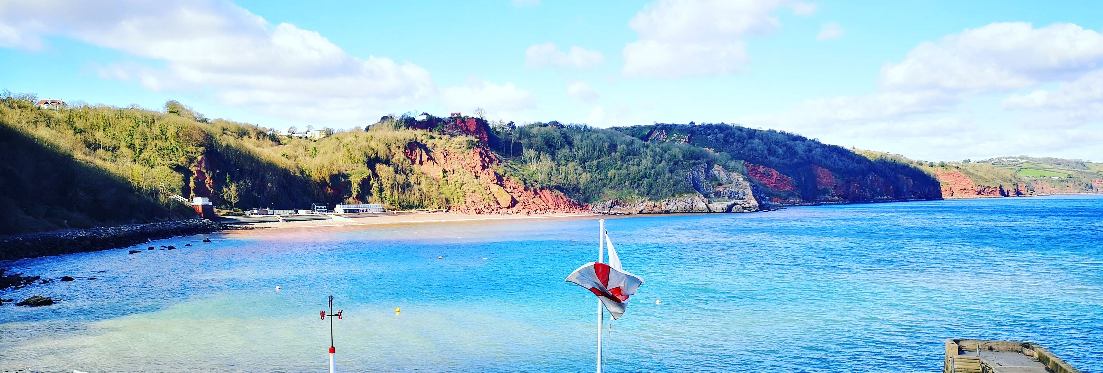 Seaside view of a sheltered bay with rocky shore, a small white terrace and tables, and wooded cliffs under a blue sky.