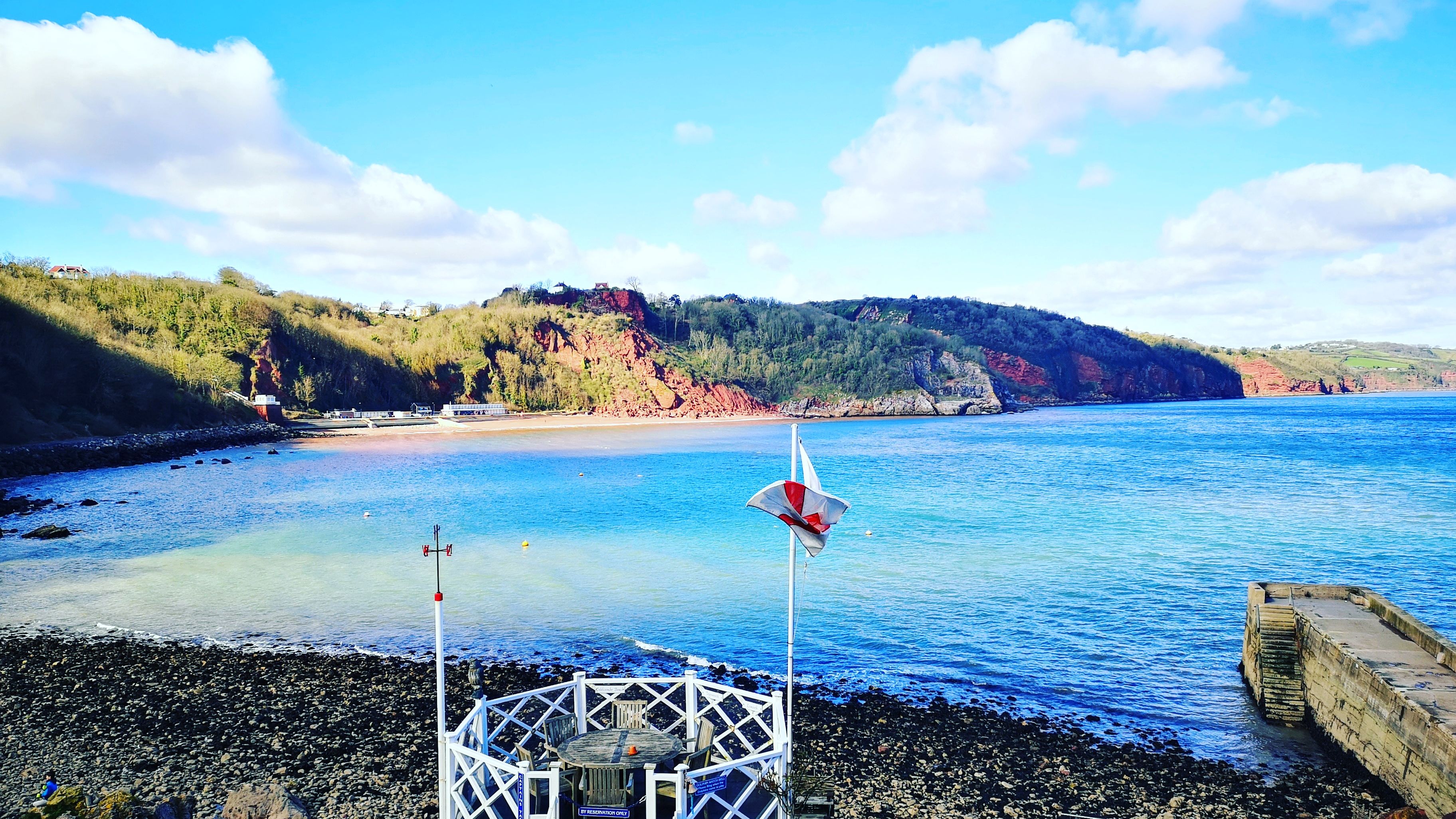 Seaside view of a sheltered bay with rocky shore, a small white terrace and tables, and wooded cliffs under a blue sky.