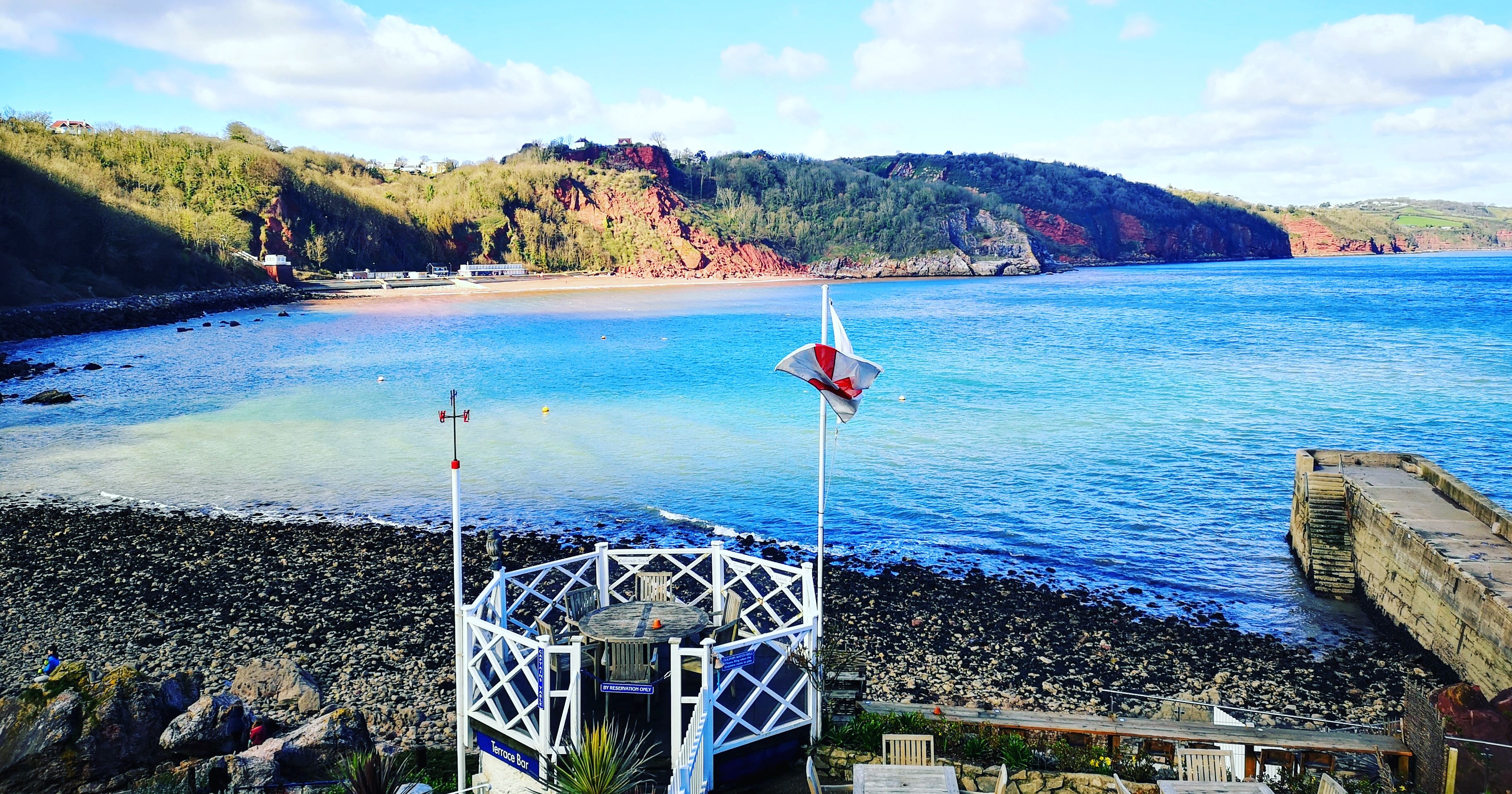 Seaside view of a sheltered bay with rocky shore, a small white terrace and tables, and wooded cliffs under a blue sky.