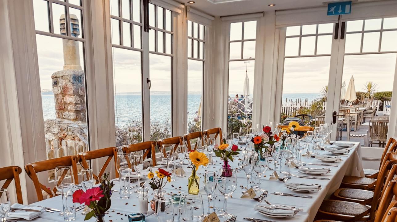 Long dining table set for a celebration with flowers and place settings by sea-view windows