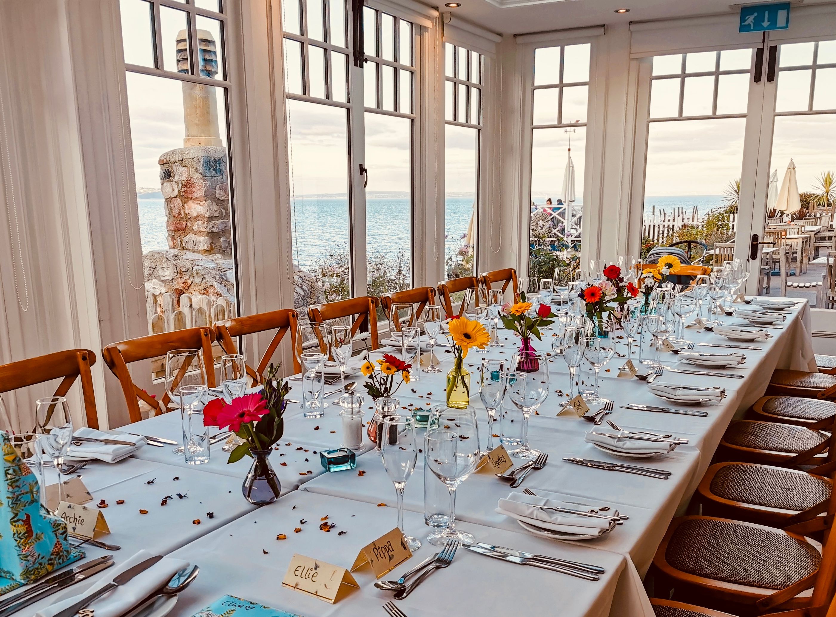 Long dining table set for a celebration with flowers and place settings by sea-view windows
