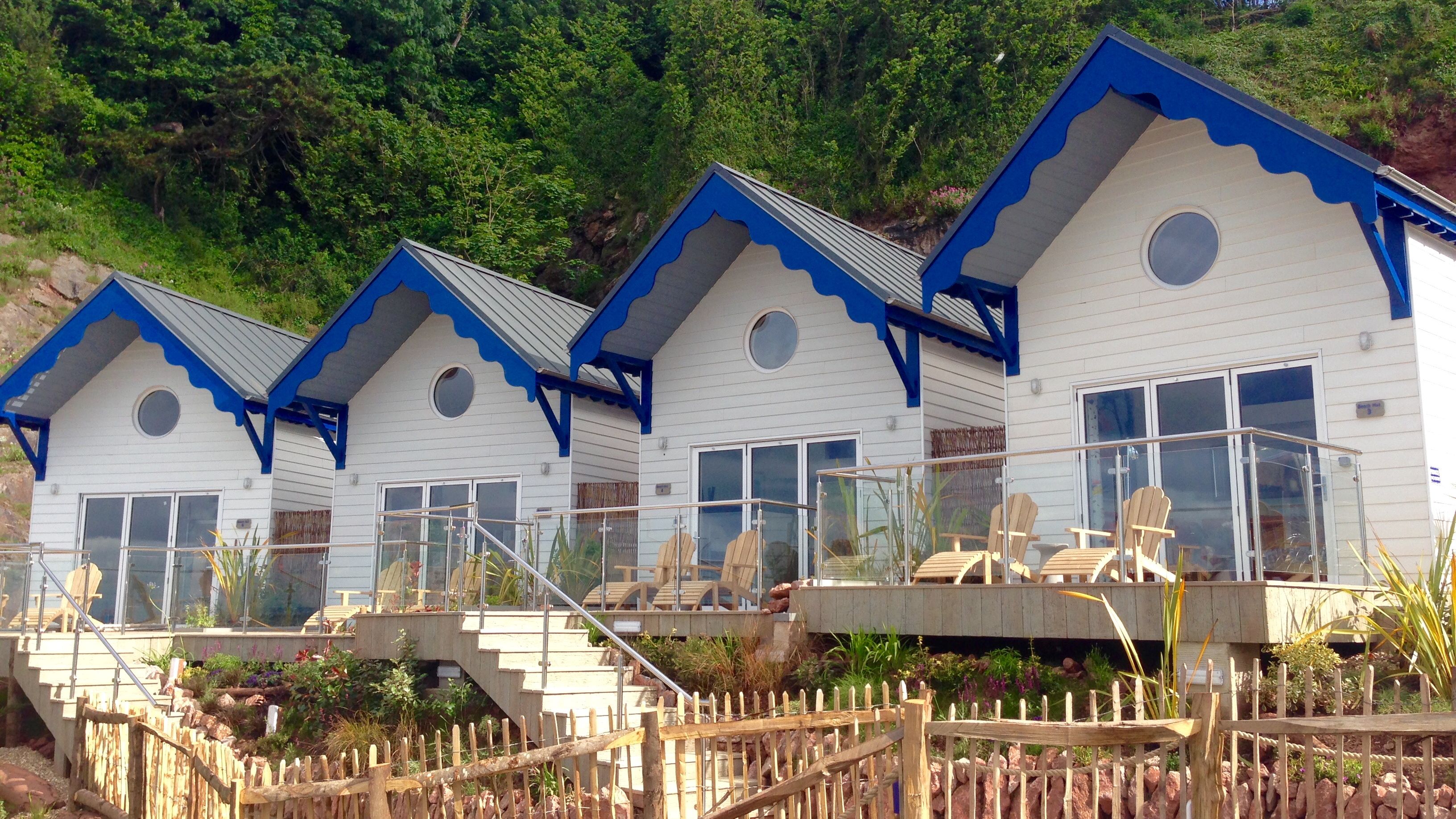 Row of white seaside cottages with blue trim and glass-railed terraces in front of a green hillside