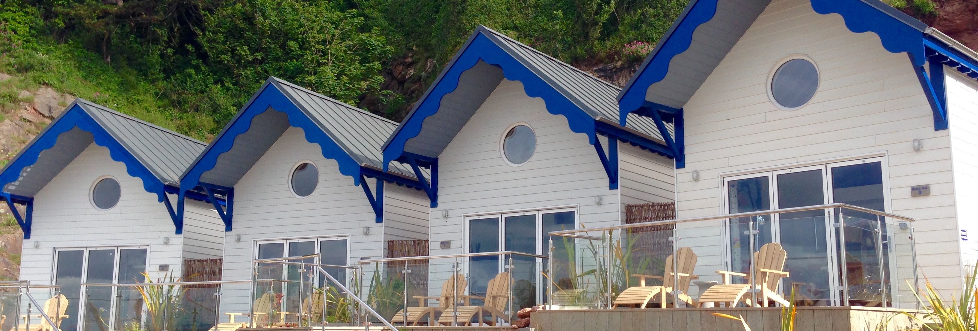 Row of white seaside cottages with blue trim and glass-railed terraces in front of a green hillside
