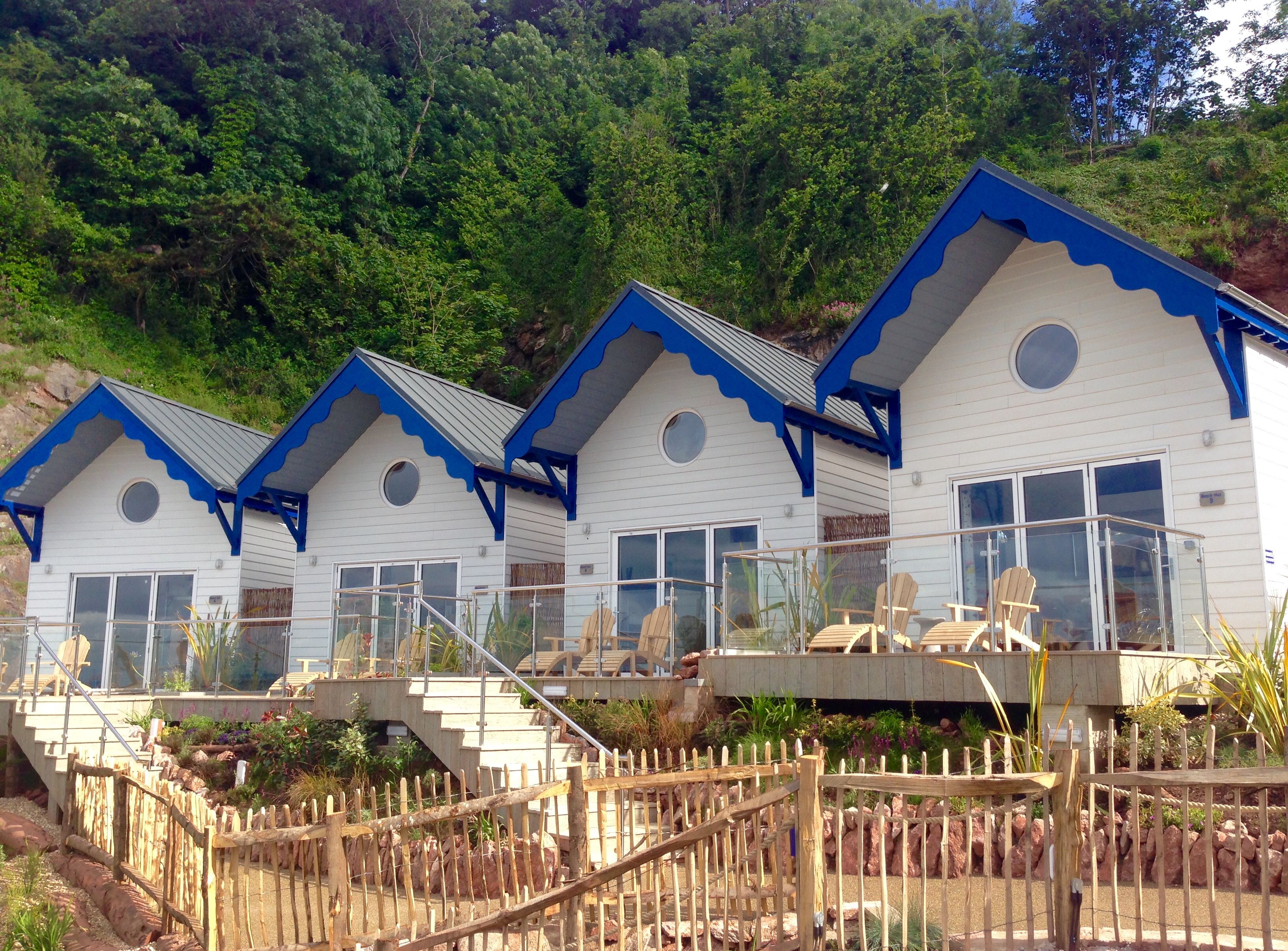 Row of white seaside cottages with blue trim and glass-railed terraces in front of a green hillside