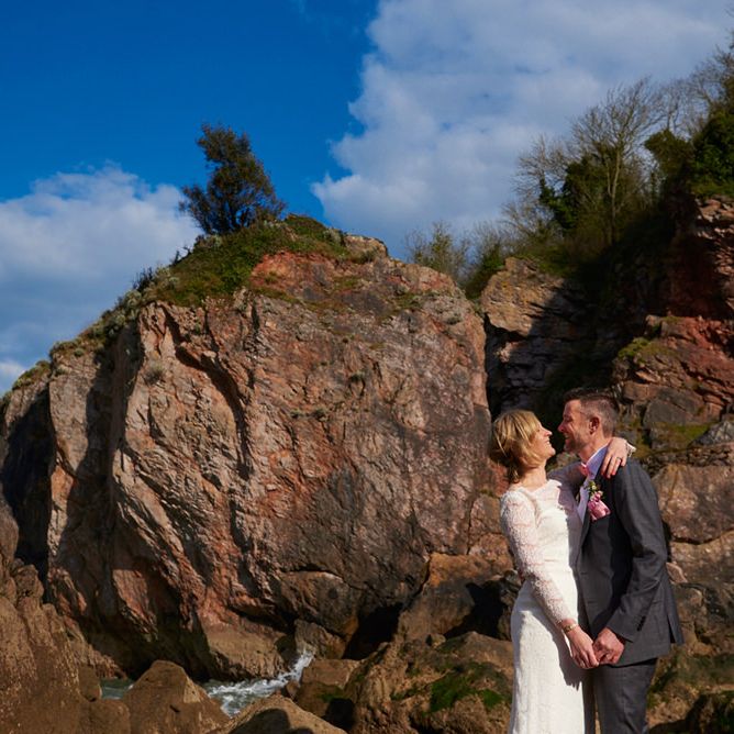 Bride and groom embracing beside rugged coastal cliffs under a blue sky in Devon