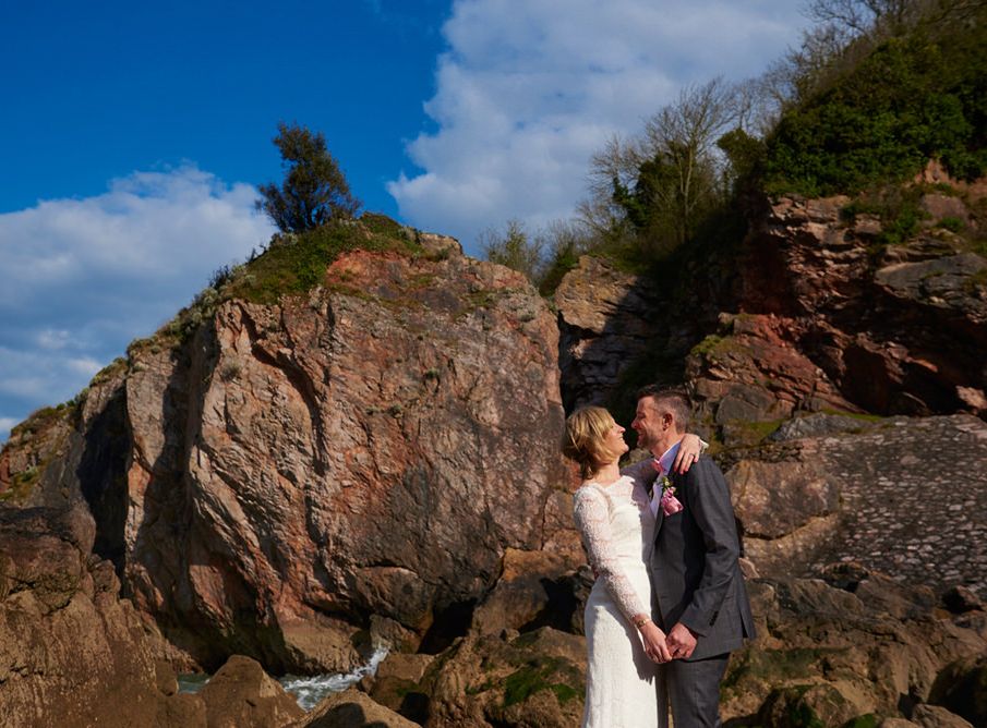Bride and groom embracing beside rugged coastal cliffs under a blue sky in Devon