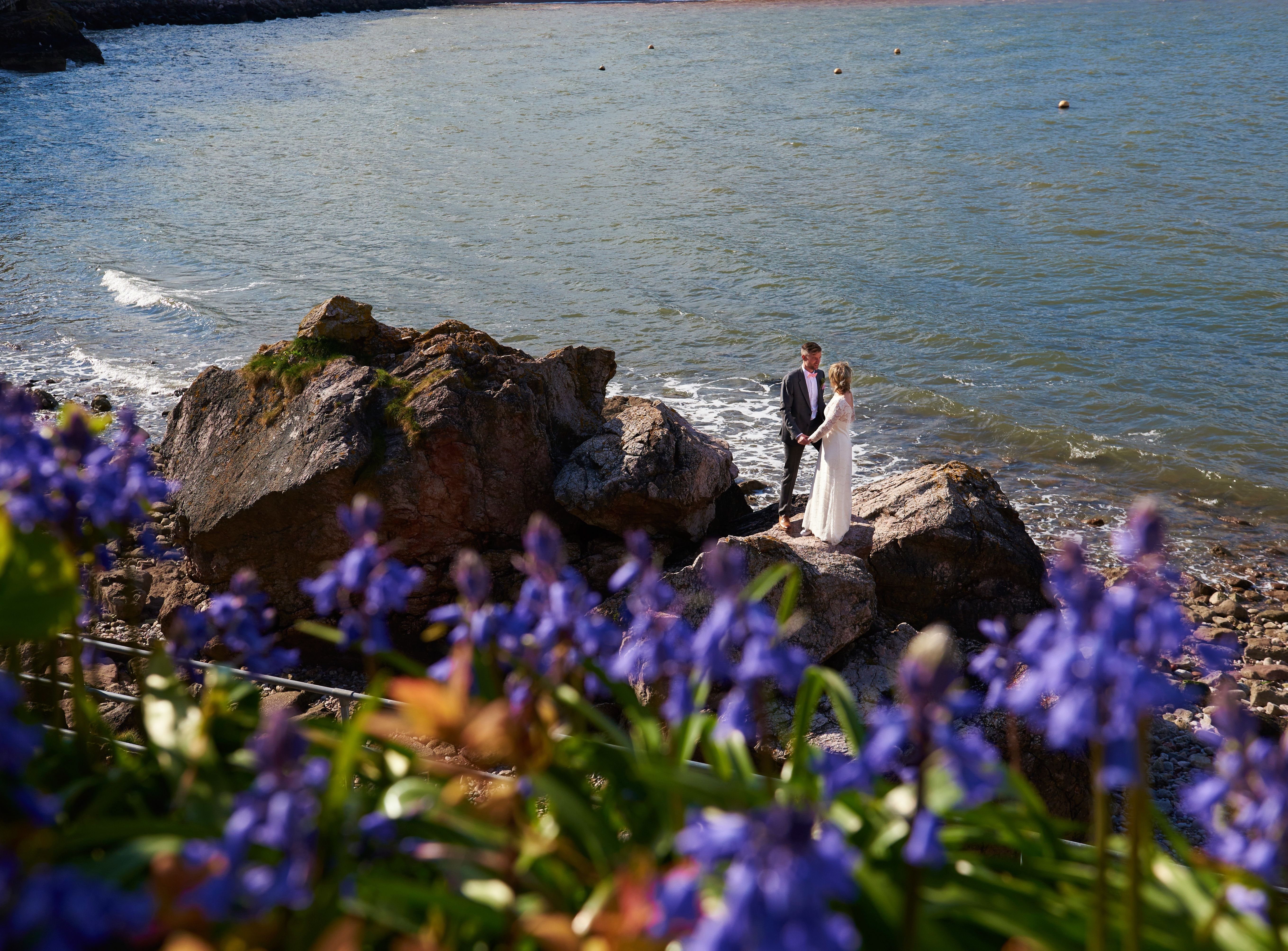 Bride and groom holding hands on rocky shoreline with sea in the background