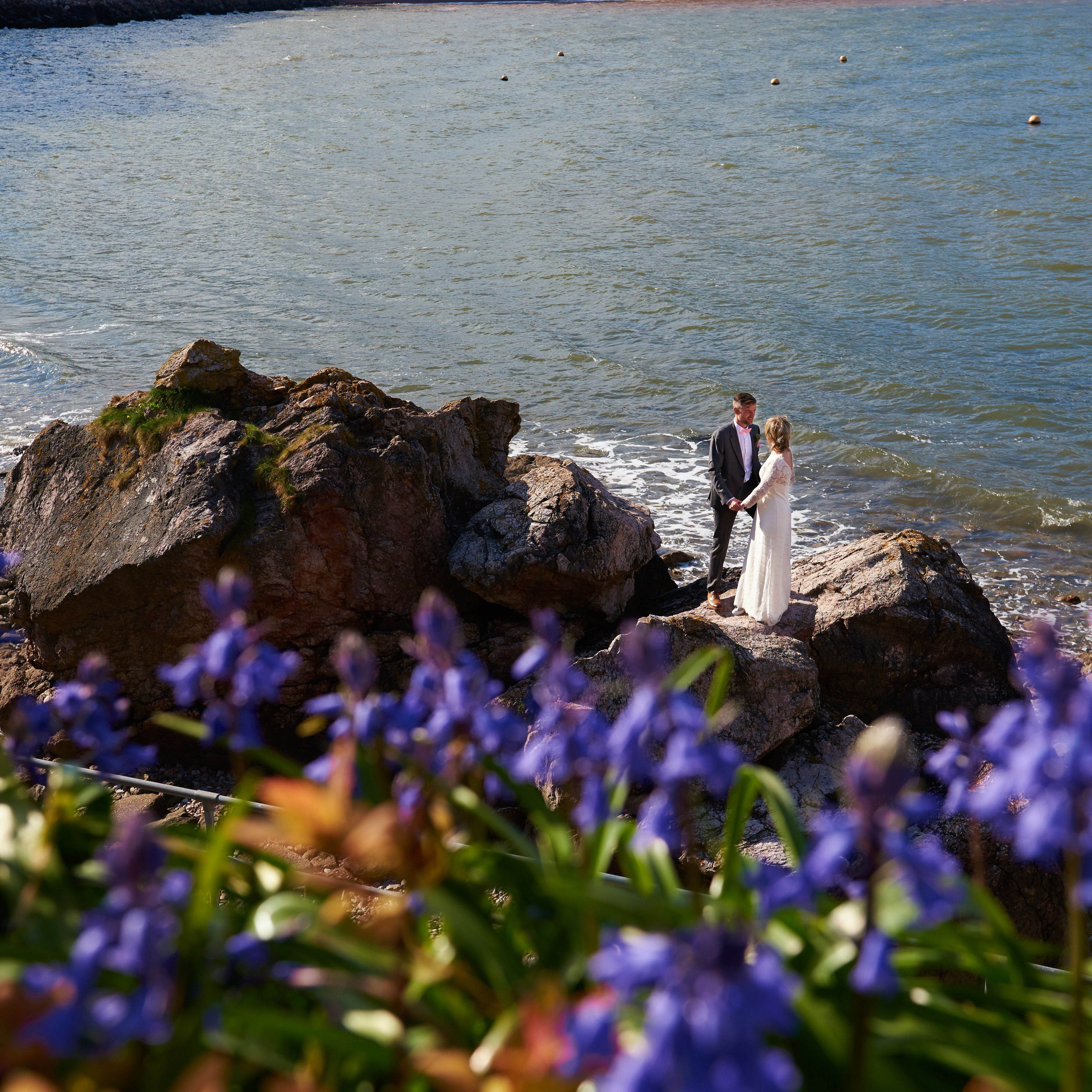 Bride and groom holding hands on rocky shoreline with sea in the background