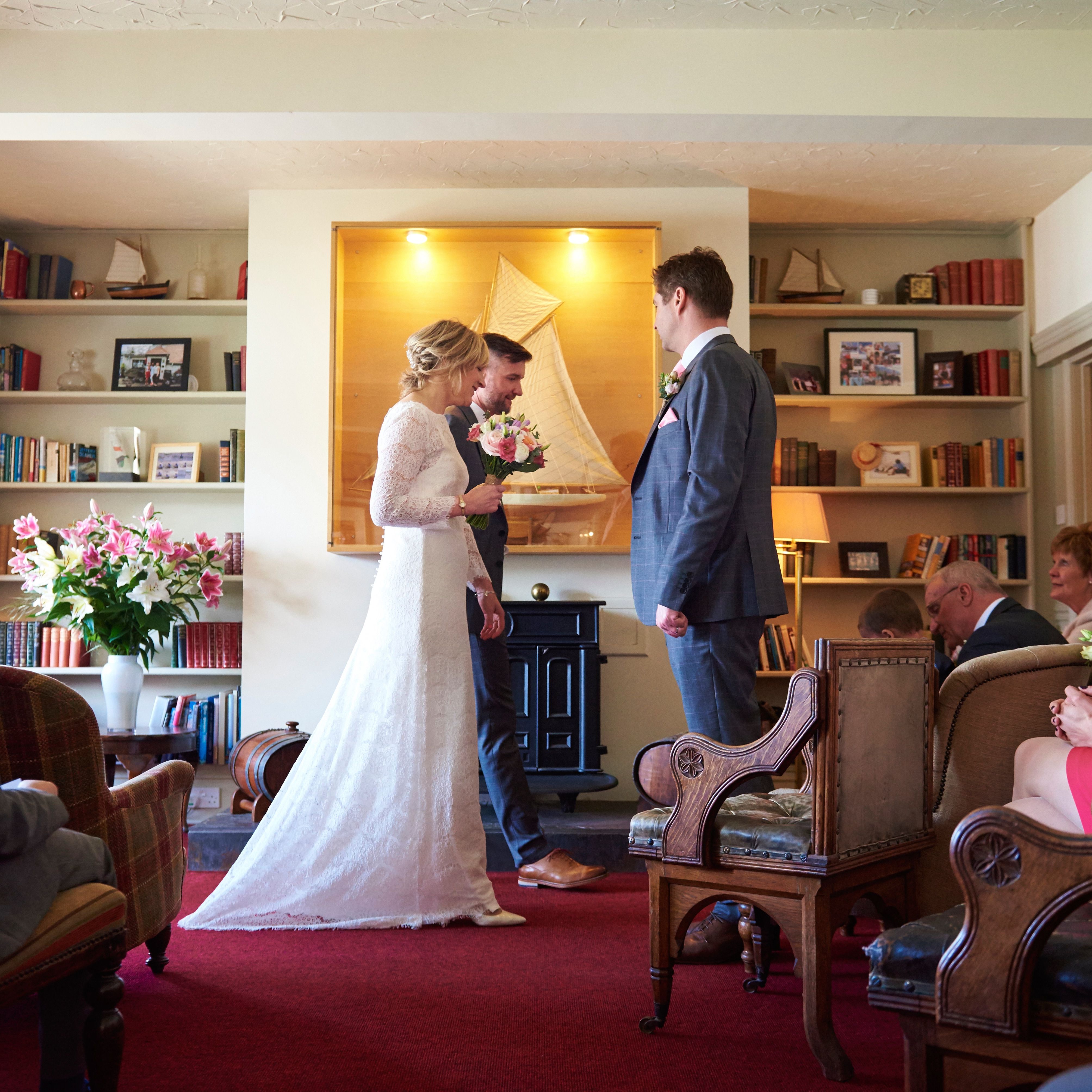 Bride holding a bouquet approaches the groom during an indoor wedding ceremony, with guests seated in a cosy room lined with bookshelves.