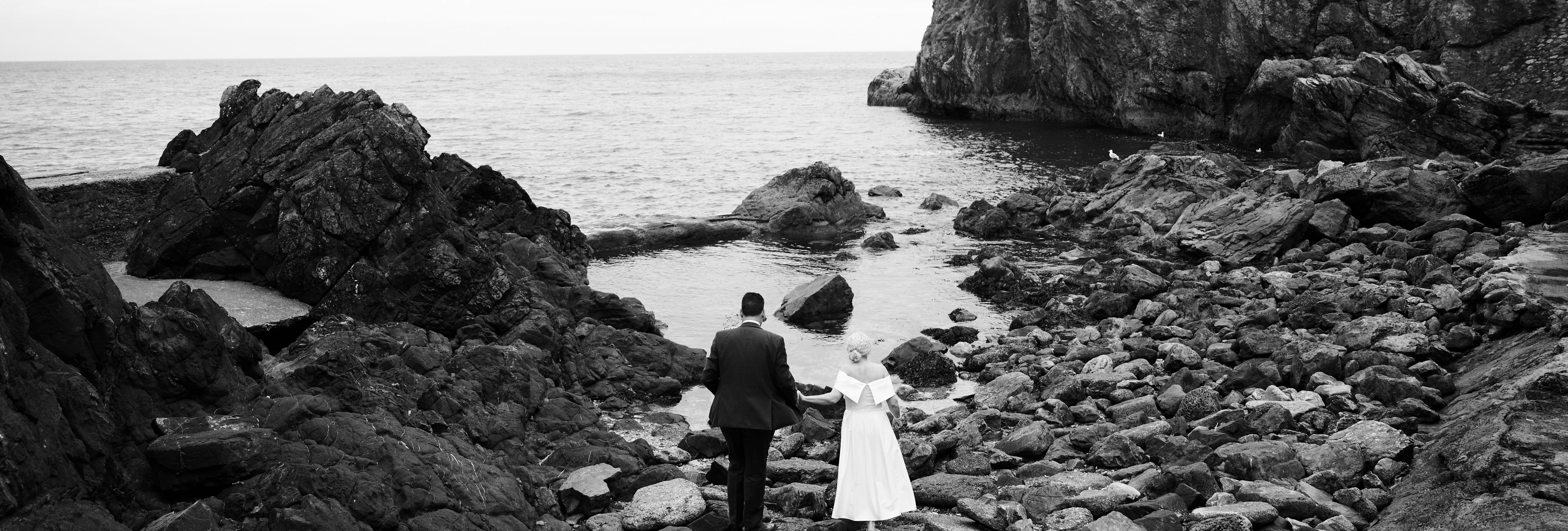 Black-and-white photo of a couple in wedding attire walking hand in hand across a rocky shoreline towards the sea