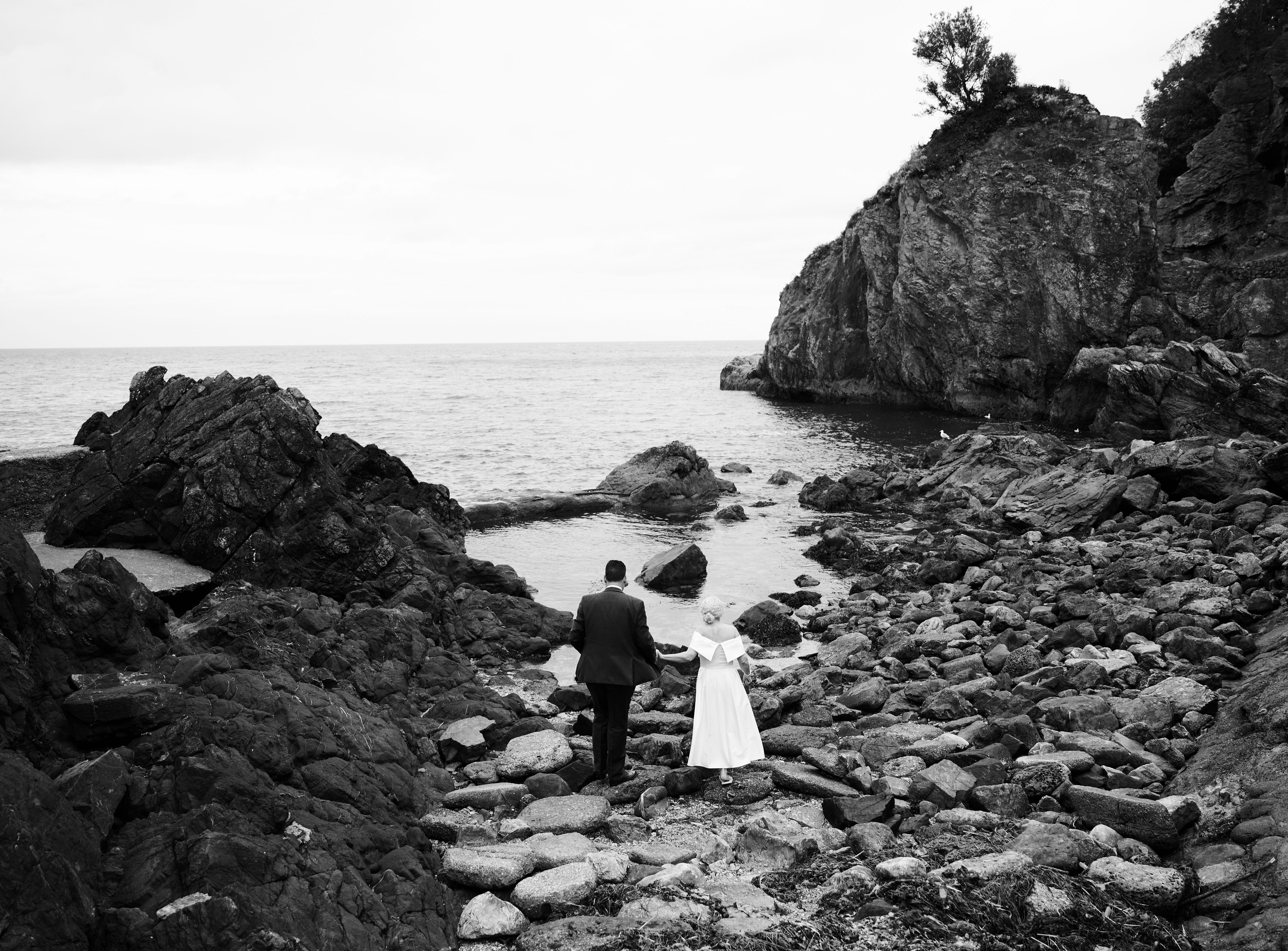 Black-and-white photo of a couple in wedding attire walking hand in hand across a rocky shoreline towards the sea
