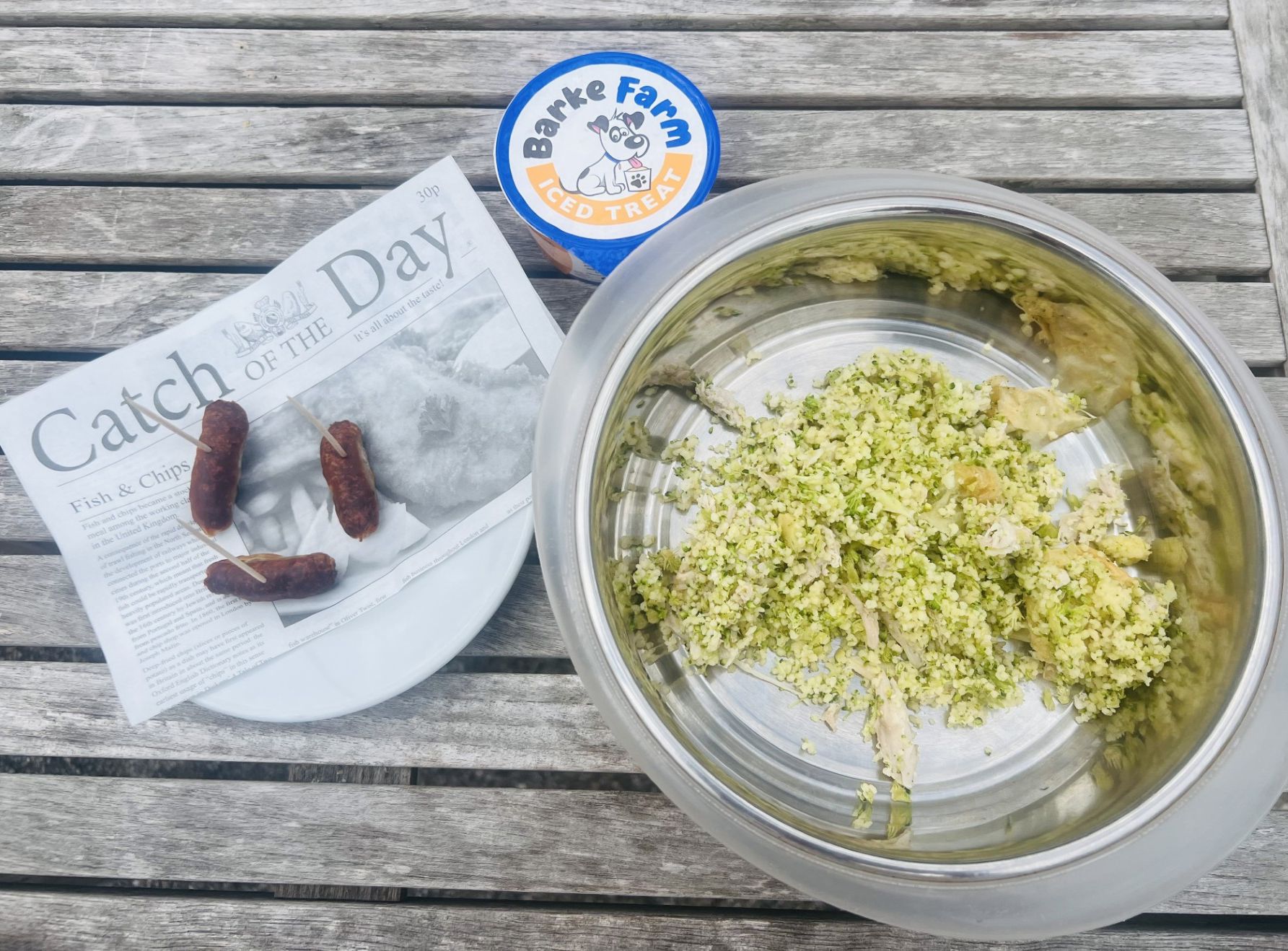 Dog food bowl with broccoli rice mix, sausages on a plate and a tub of Barke Farm iced treat on a wooden table