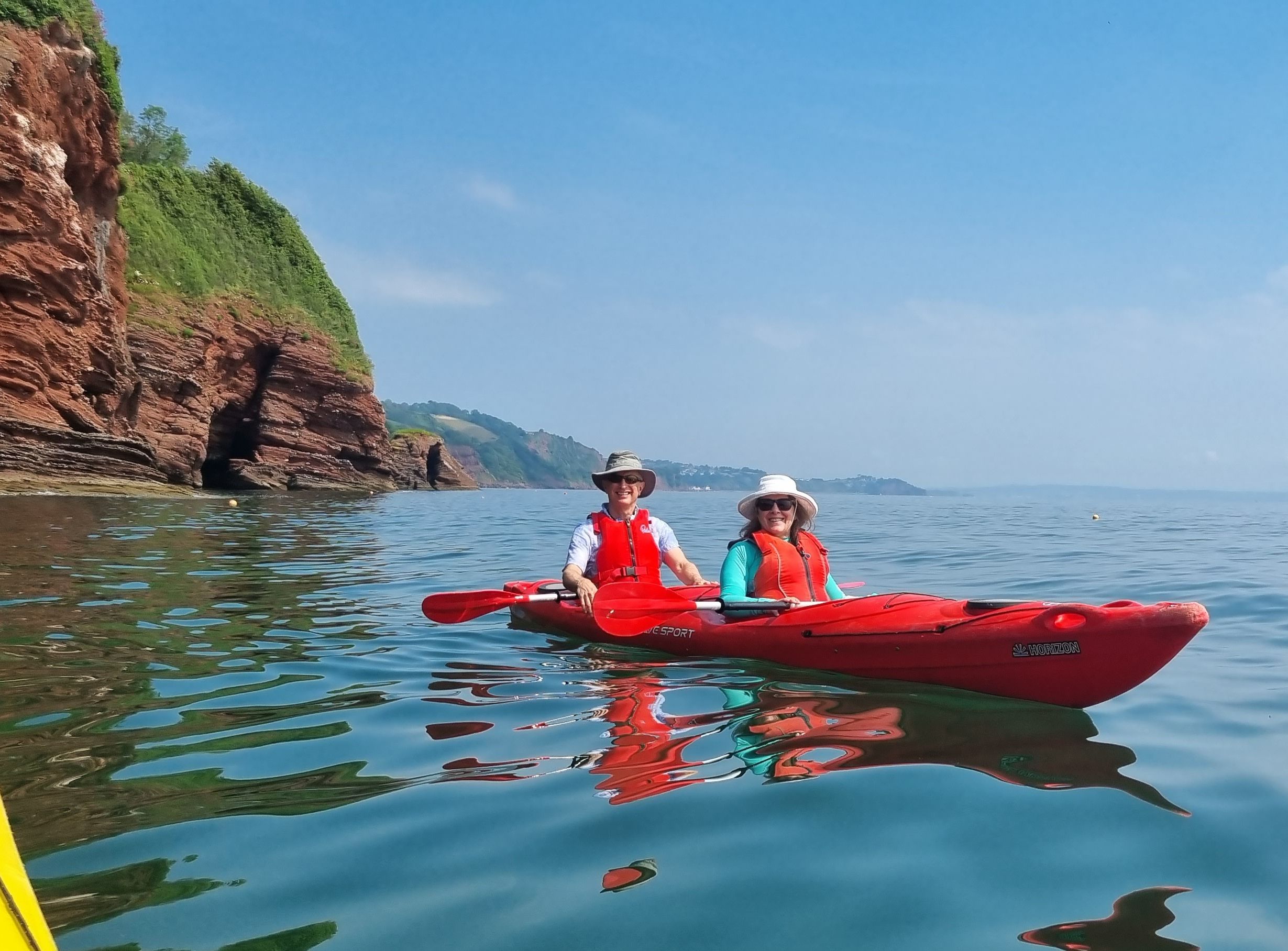 Two people kayaking in a red tandem kayak on calm sea beside red cliffs
