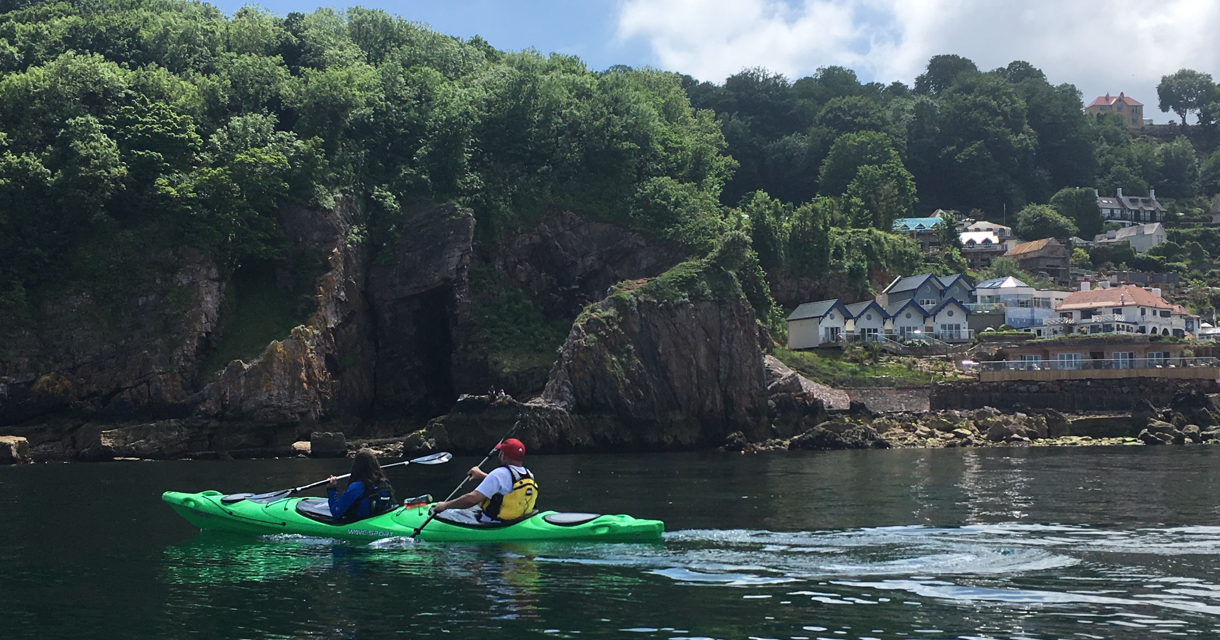 Two people kayaking in a green kayak on calm water near a rocky, tree-covered coastline with seaside houses.