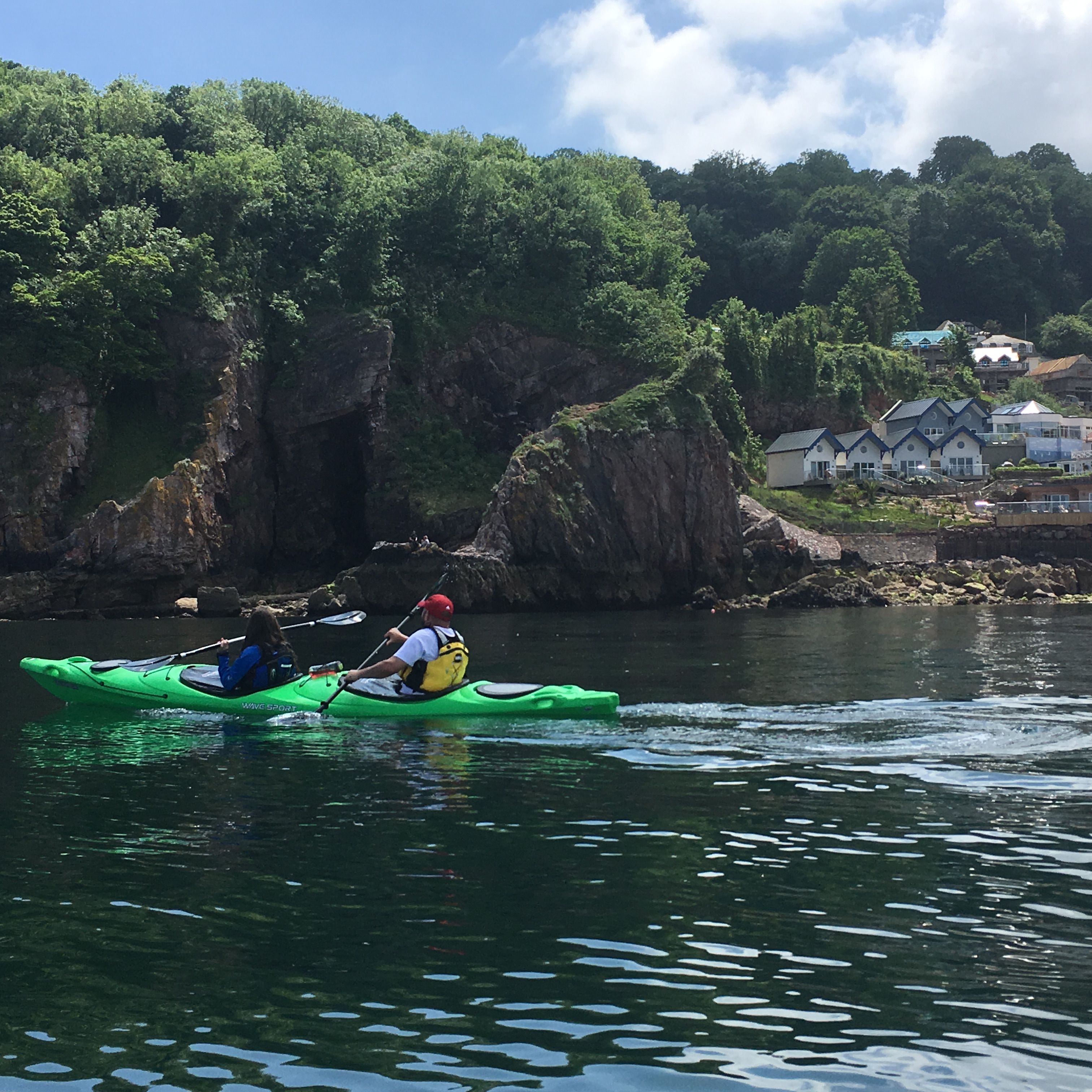 Two people kayaking in a green kayak on calm water near a rocky, tree-covered coastline with seaside houses.