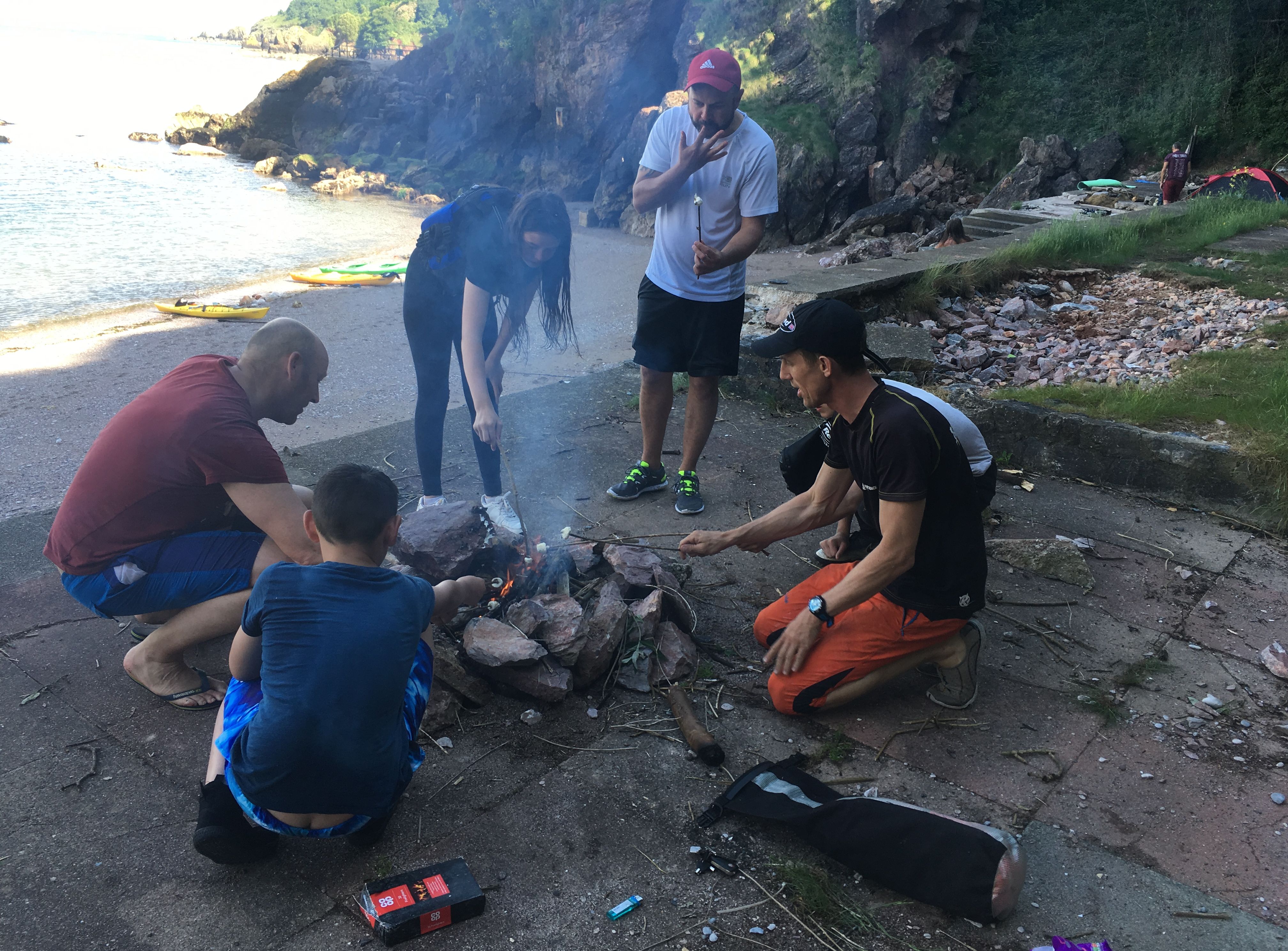 Group of friends around a small campfire on a rocky beach, roasting marshmallows beside the sea