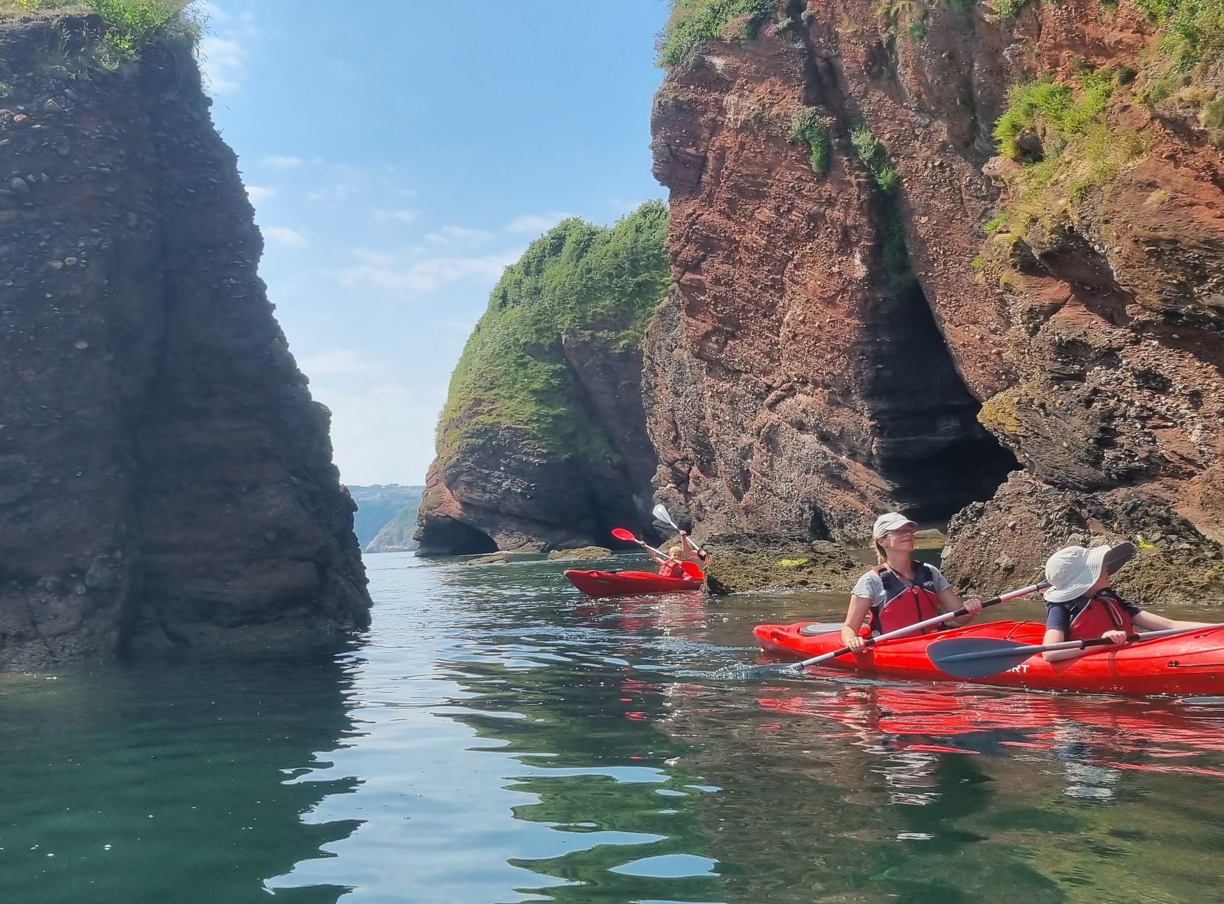 Two people kayaking in red boats beside rugged sea cliffs and a natural rock arch