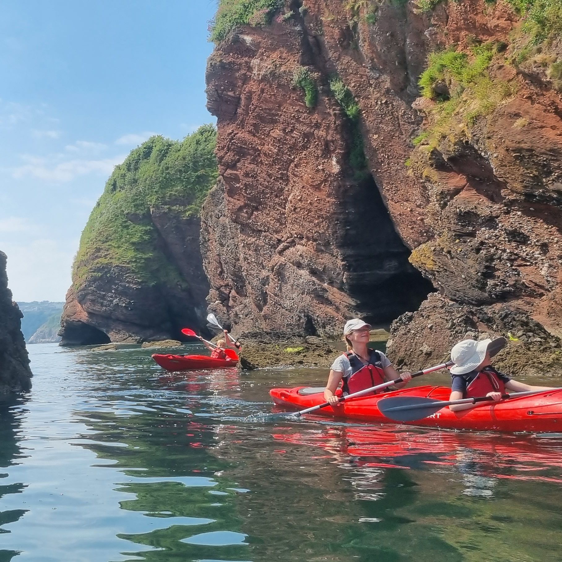 Two people kayaking in red boats beside rugged sea cliffs and a natural rock arch