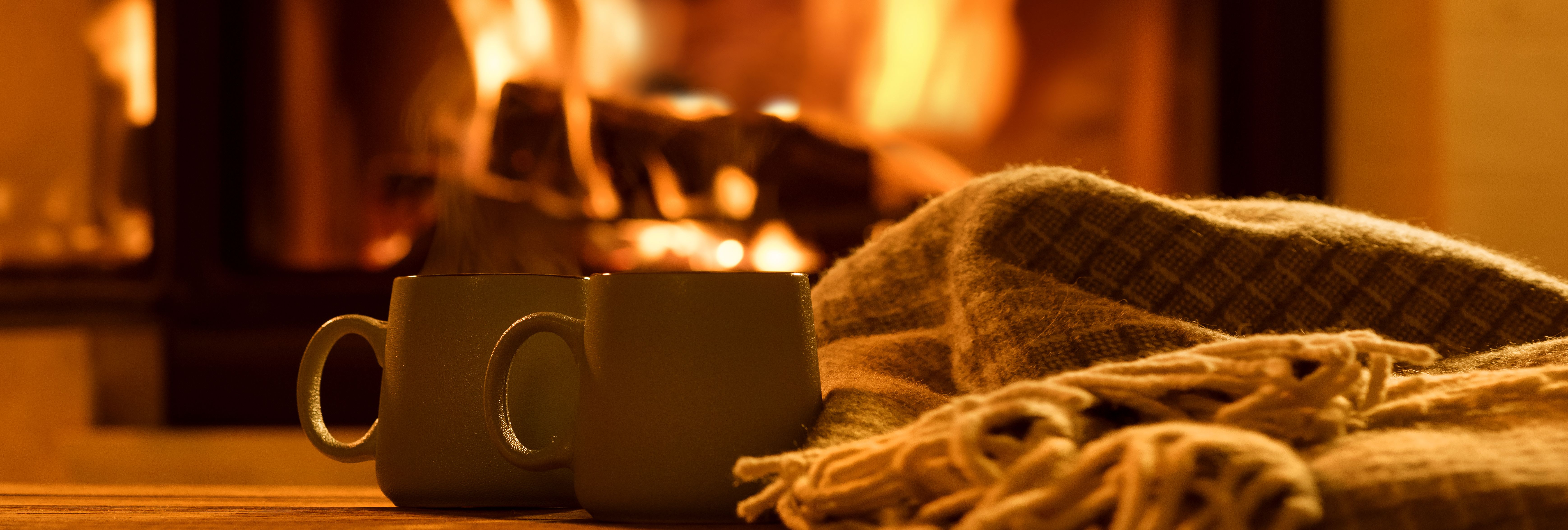 Two mugs on a wooden table beside a woollen blanket, with a roaring fireplace in the background