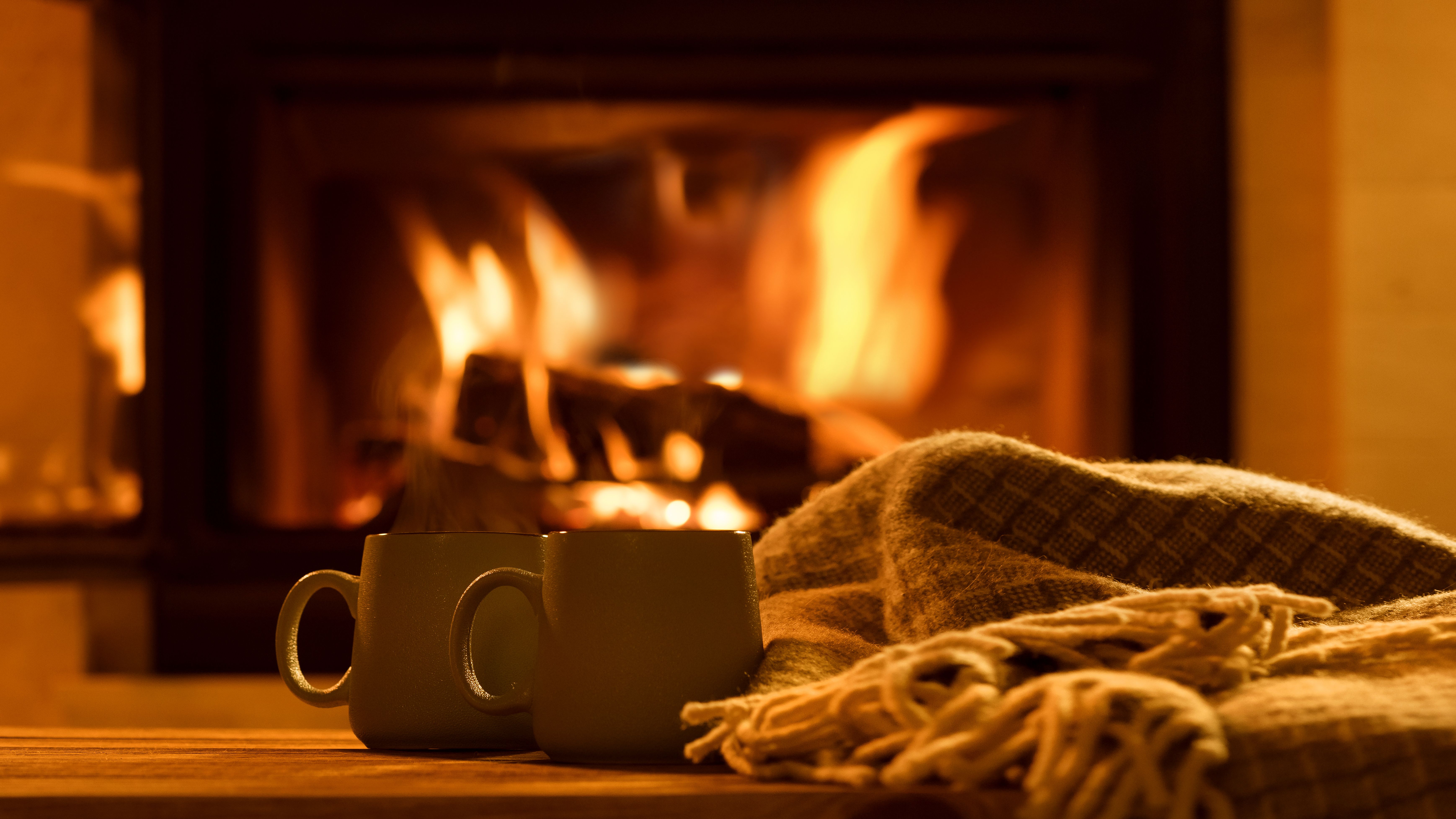 Two mugs on a wooden table beside a woollen blanket, with a roaring fireplace in the background
