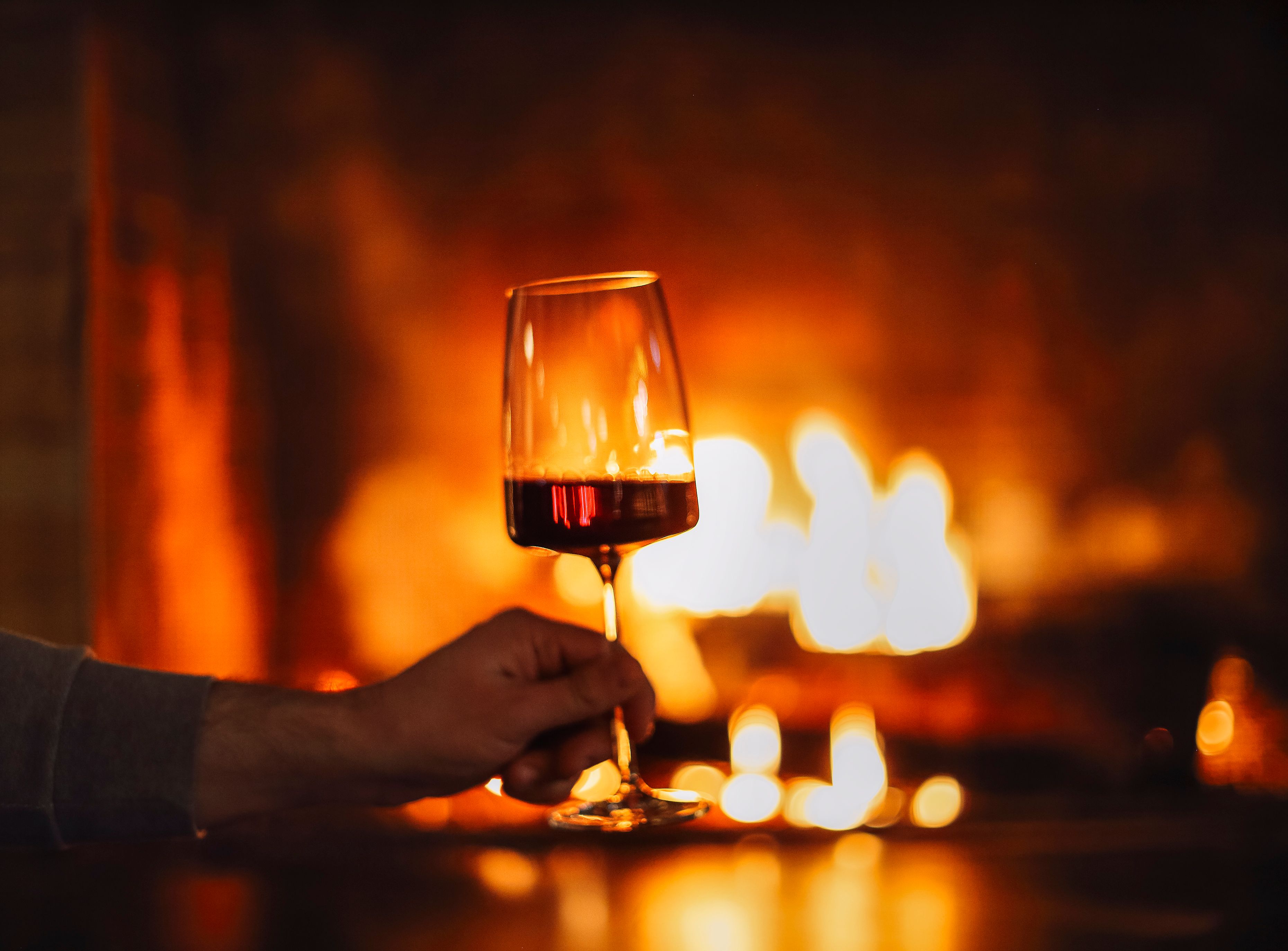Hand holding a glass of red wine in front of a glowing fireplace